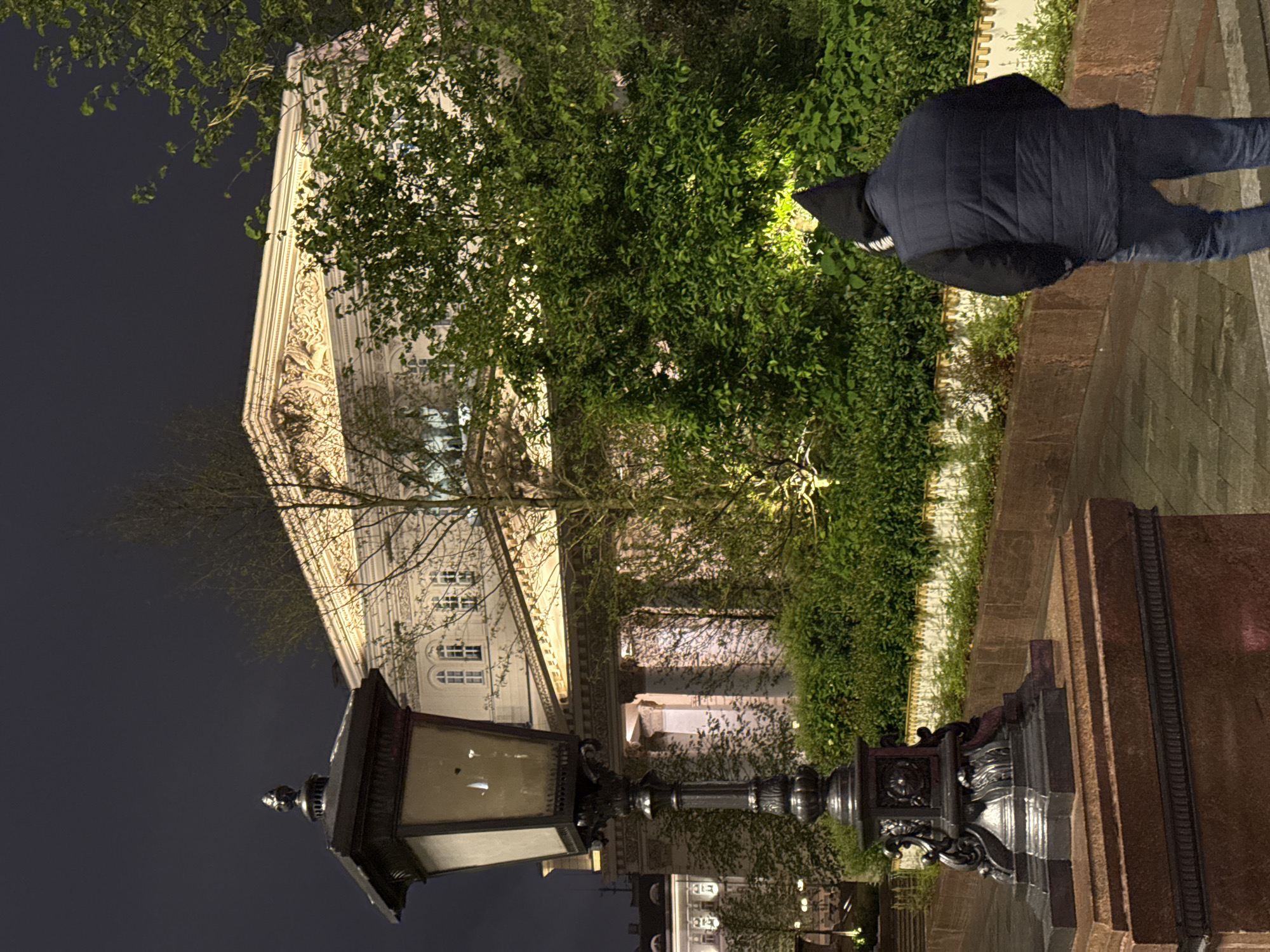 At night in Moscow, Russia, a hooded figure walks along a sidewalk next to a building adorned with decorative carvings. A vintage lamppost stands in the foreground. The building's exterior is lit, and a large, leafy tree grows in front of it. Further down the street, other buildings with illuminated windows are visible. The sidewalk is paved with stone.