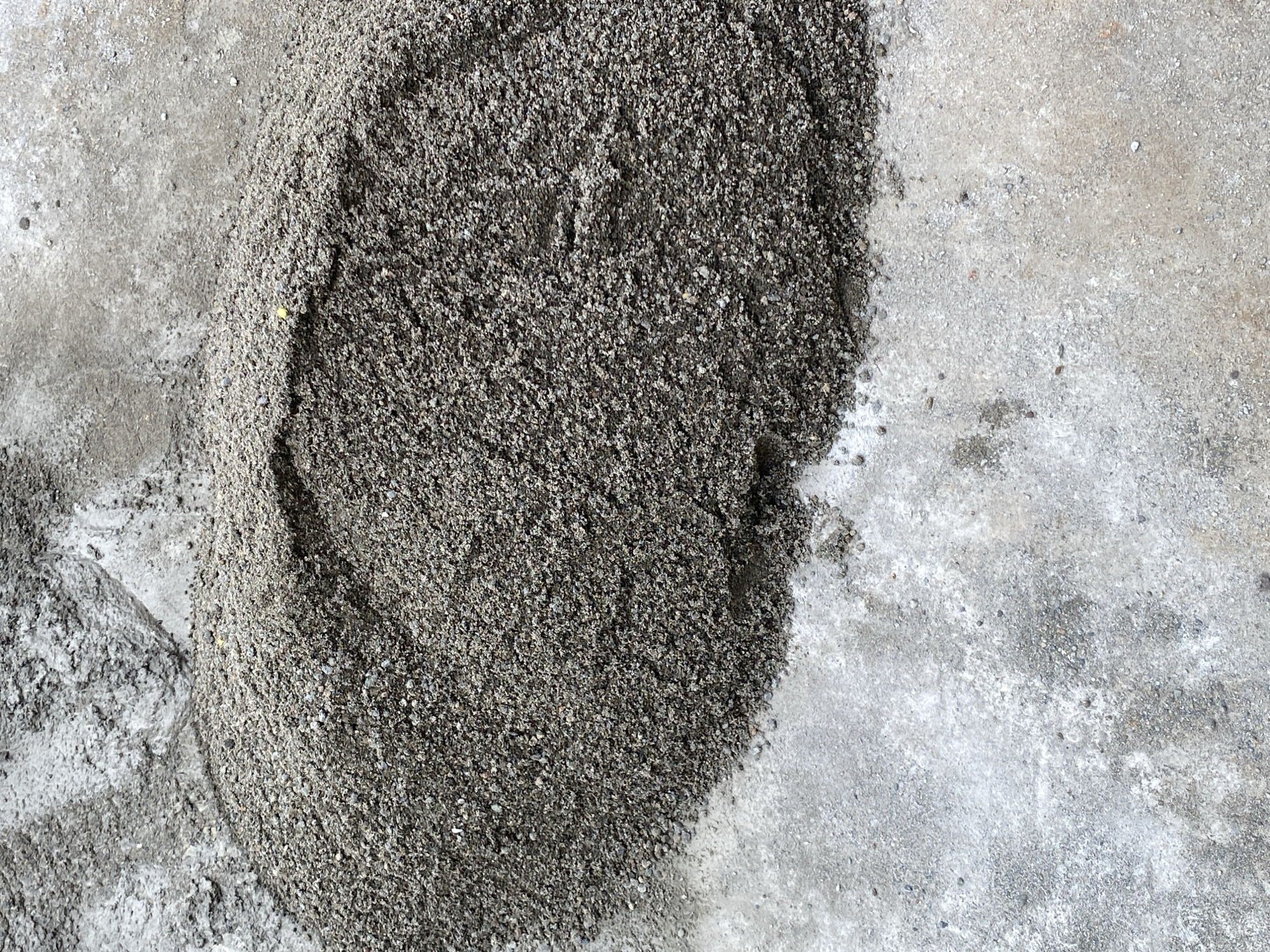 A pile of dark, granular material, possibly sand or gravel, sits on a light-colored, rough surface, likely concrete. The pile is unevenly shaped and appears to be a construction material. The background shows more of the rough, concrete-like surface with some lighter, powdery residue. The lighting suggests an outdoor, possibly sunny, environment. No people are visible in the image.