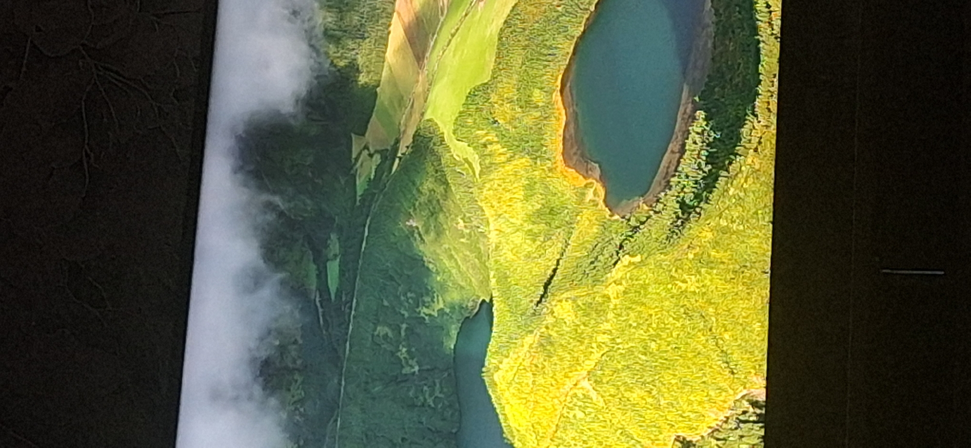 The image is an aerial view of a lush green landscape featuring several bodies of water, likely lakes or volcanic craters, surrounded by dense vegetation. The terrain is undulating, with hills covered in trees and patches of lighter green fields. Wisps of fog or clouds are visible, particularly in the upper portion of the image, obscuring some of the distant landscape. The bodies of water have a deep blue-green hue. The overall scene depicts a natural, possibly remote, and scenic environment. There is no visible indication of the city of Bila Tserkva, Ukraine, or any man-made structures that would identify it as such.