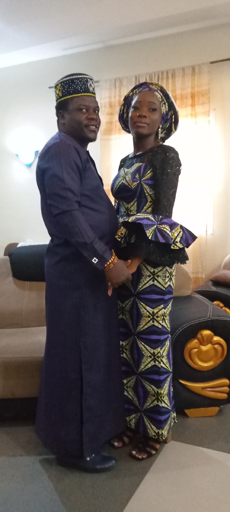A photo shows a man and a woman standing indoors, holding hands. The man is wearing a long-sleeved, dark blue robe, a beaded bracelet, and a dark hat with gold and white details. The woman is wearing a patterned dress with a black lace top and a headwrap of similar fabric. A sofa and a black and gold object are visible in the background. The location is Unknown, Unknown.