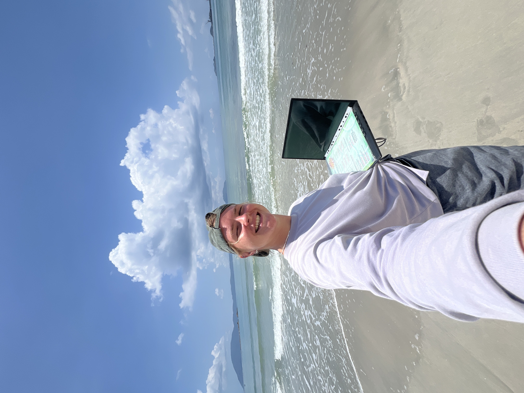 A person is seen holding a laptop and documentation while standing on a beach in Dong Ninh Hoa Ward, Vietnam. They are facing the camera and smiling. The beach is sandy, and the ocean is visible in the background. The sky is blue with some clouds.