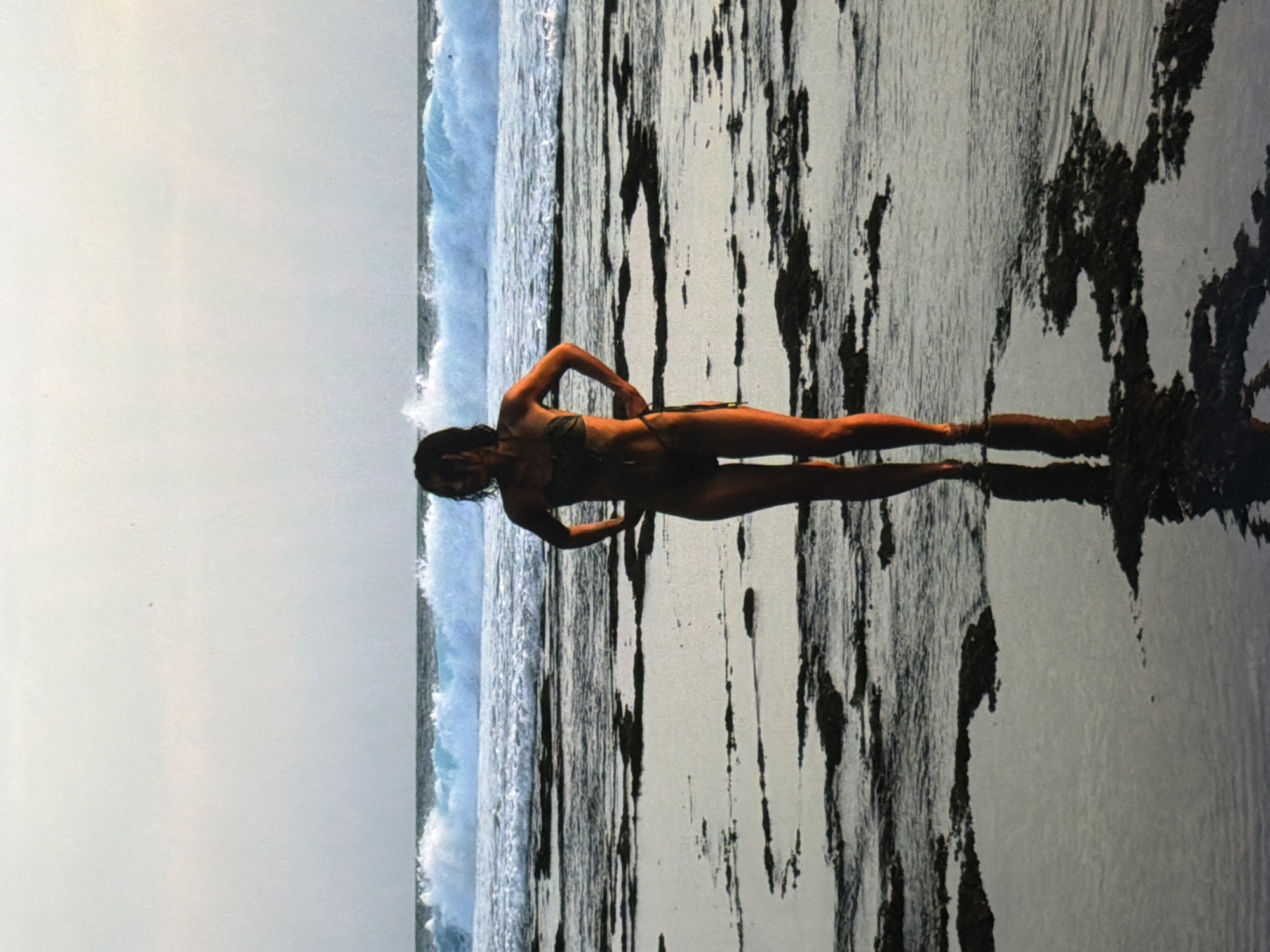 A woman in a dark-colored bikini stands on a textured, rocky surface near the ocean in what appears to be North Kuta, Indonesia. The water is visible in the background, with waves breaking. The sky is overcast. The woman's body is positioned horizontally in the frame, and she is striking a pose with her hands on her hips. The textured surface on which she is standing consists of a light-colored base with dark, irregular markings.