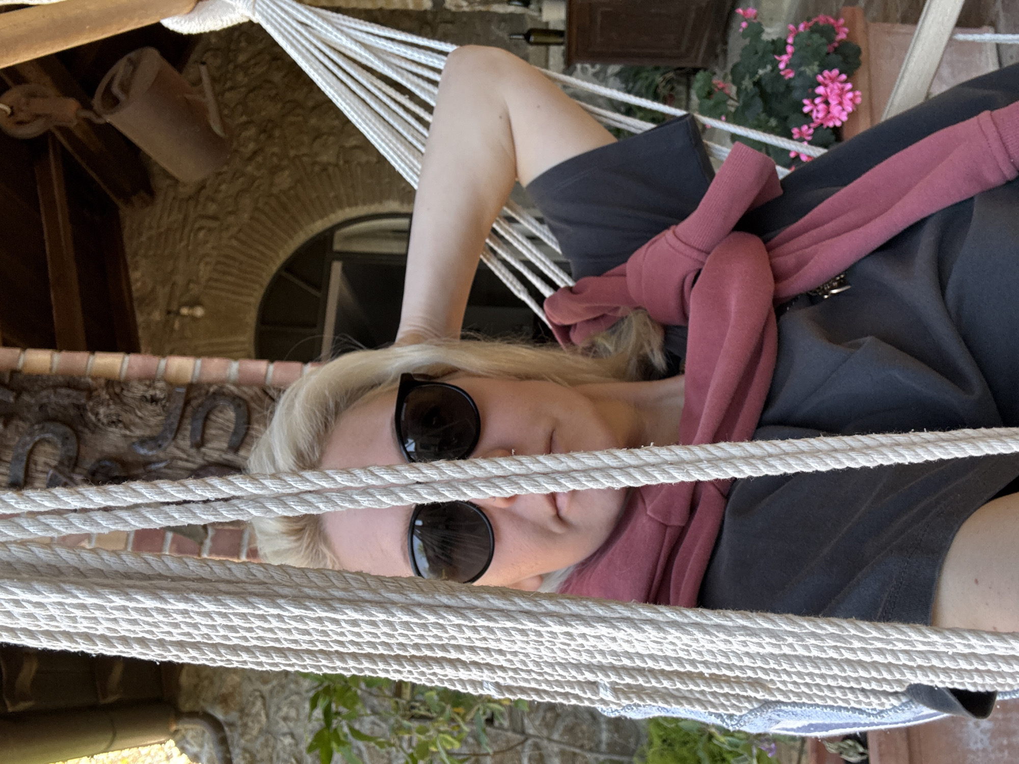 A fair-skinned person wearing sunglasses is reclining in a white rope hammock. The person has blond hair and a nose ring. The person is wearing a dark-colored t-shirt and a red sweatshirt tied around their neck.
Behind the hammock, there is a stone archway. To the left of the archway, there is a decorative metal piece with horseshoe shapes. To the right of the archway, there are pink flowers in a pot. A brown bucket hangs from the ceiling in the background.
The photo was taken in Velletri, Italy.