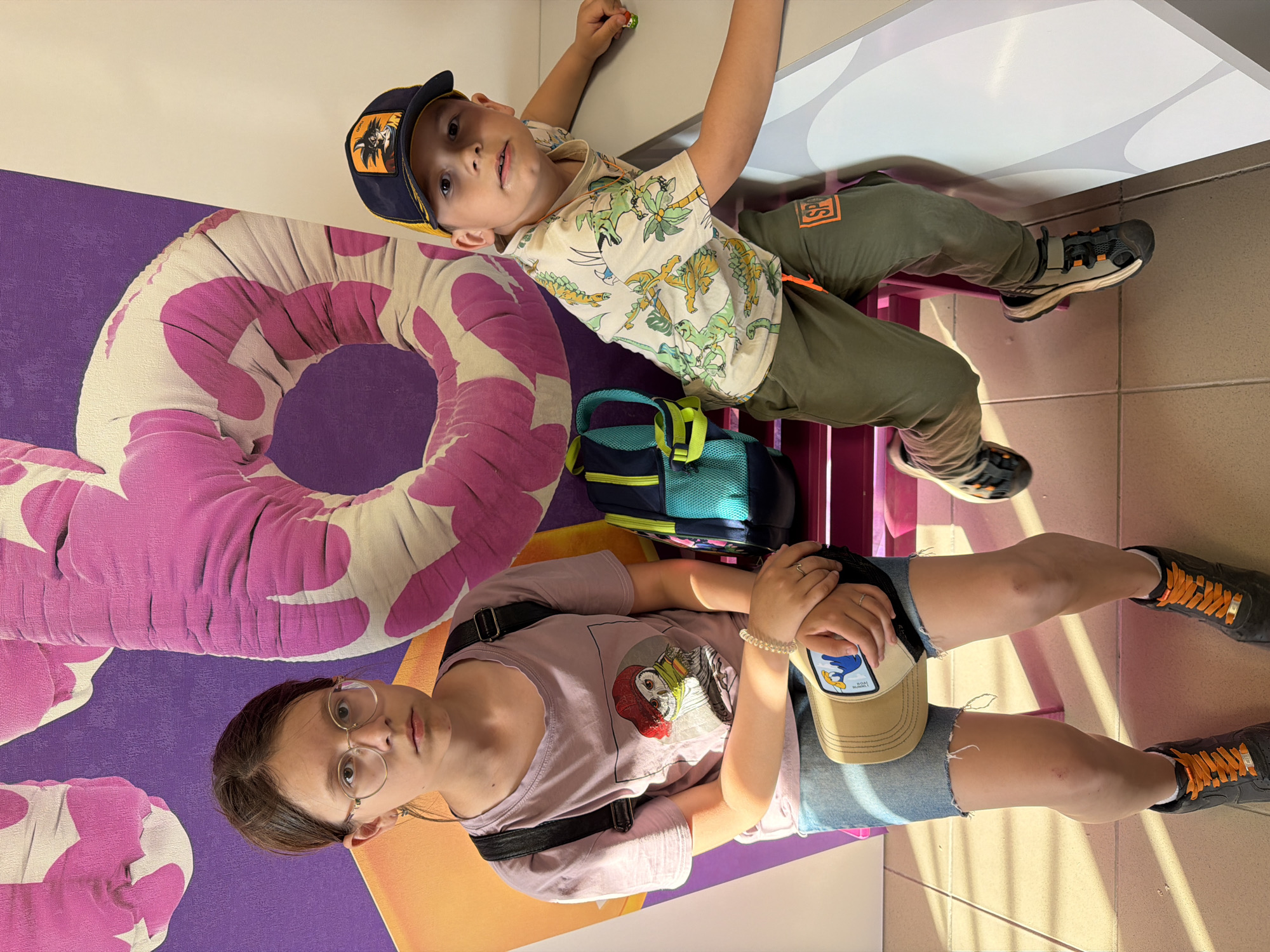 Two individuals are positioned in what appears to be an interior setting in the city of Chaikovsky, Russia.
The first individual is a young boy, wearing a blue and yellow baseball cap featuring an anime character, a short-sleeved shirt with a dinosaur print, and olive green pants with an orange square. He is lying on a platform.
The second individual is a teenage girl wearing glasses, a lavender-colored t-shirt with a cartoon character printed on it, and denim shorts. She also has a tan baseball cap in her hands, resting on her legs. She is wearing a black cross-body bag.
Both individuals are wearing dark-colored sneakers with orange laces.
The background contains a large, colorful graphic featuring a pink and white donut design on a purple surface. A teal and black backpack rests next to the boy. The setting appears to be a brightly lit, possibly public space with tiled flooring.