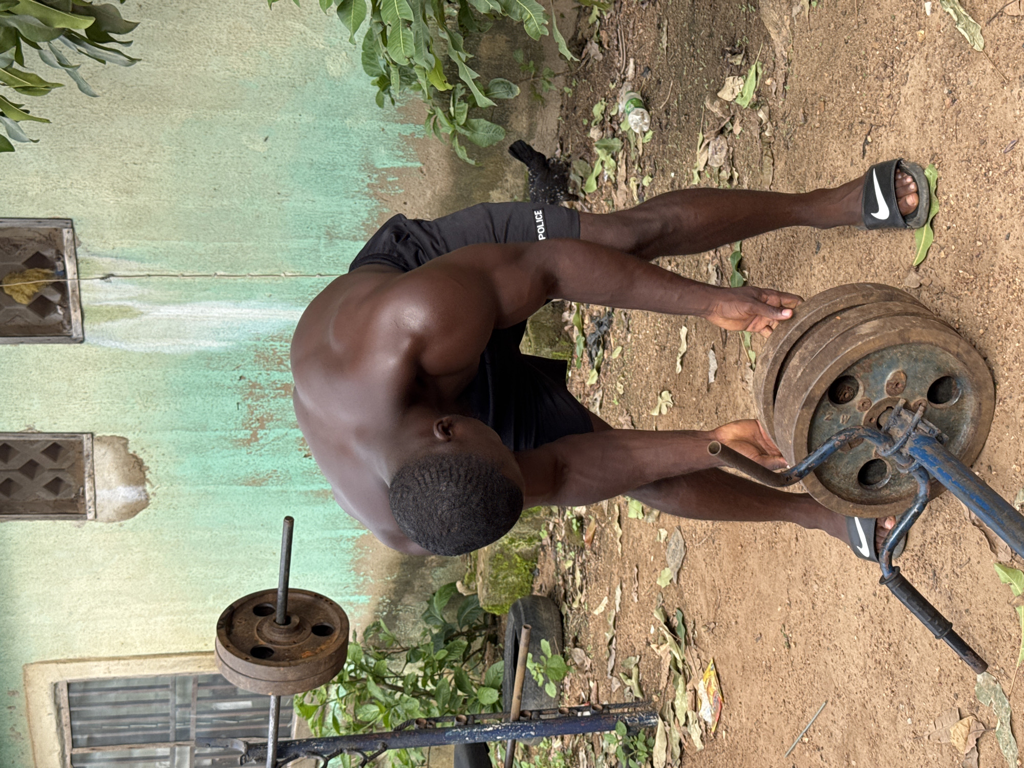 A shirtless Black man is bent over to pick up a weight plate from the ground in an outdoor setting. He is wearing black shorts with "POLICE" written on the left leg and black and white Nike slides. The ground is covered in dirt and scattered leaves. To the left of the man, a weightlifting bar is resting on a rusty blue rack, with weight plates on it. Behind the man is a faded green wall with two small, barred windows. There are some green plants and a black chicken in the background. The scene appears to be in a yard in Akwanga, Nigeria.