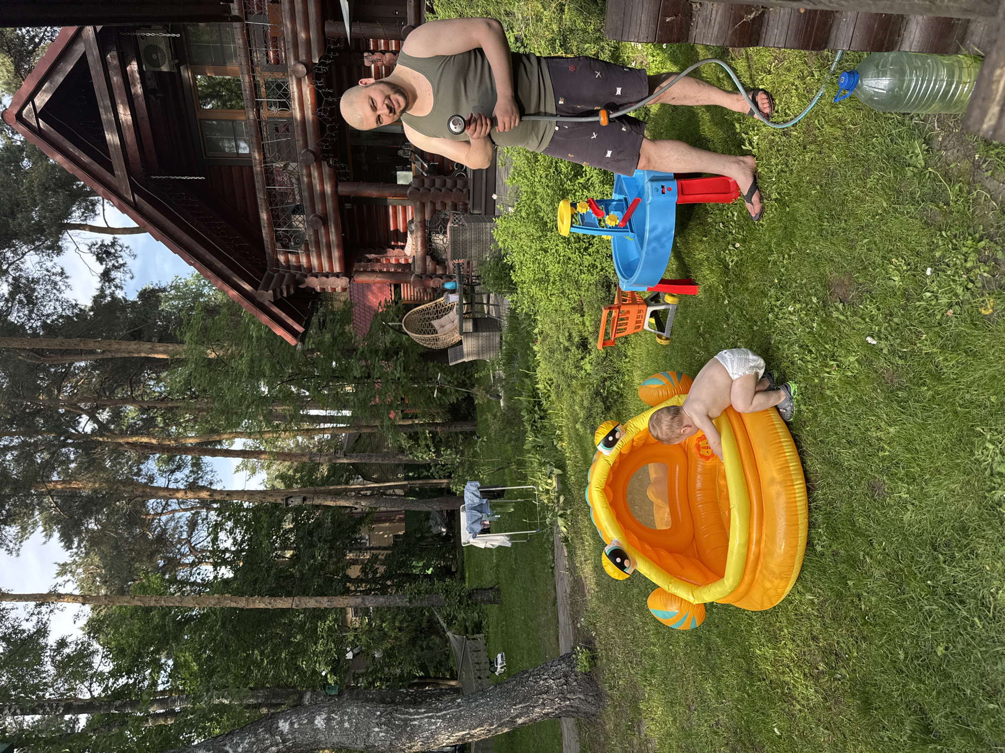 The image depicts a backyard scene in Tomilino, Russia. A bald man wearing a green tank top and dark shorts stands on a wooden deck, holding a garden hose. Next to him is a large plastic water bottle connected to the hose. In the yard below, a toddler wearing a diaper is leaning over the edge of an orange and yellow inflatable pool. Nearby, there's a blue and yellow water table and other children's toys. The background shows a log cabin-style house, trees, and a clothesline with laundry hanging on it.