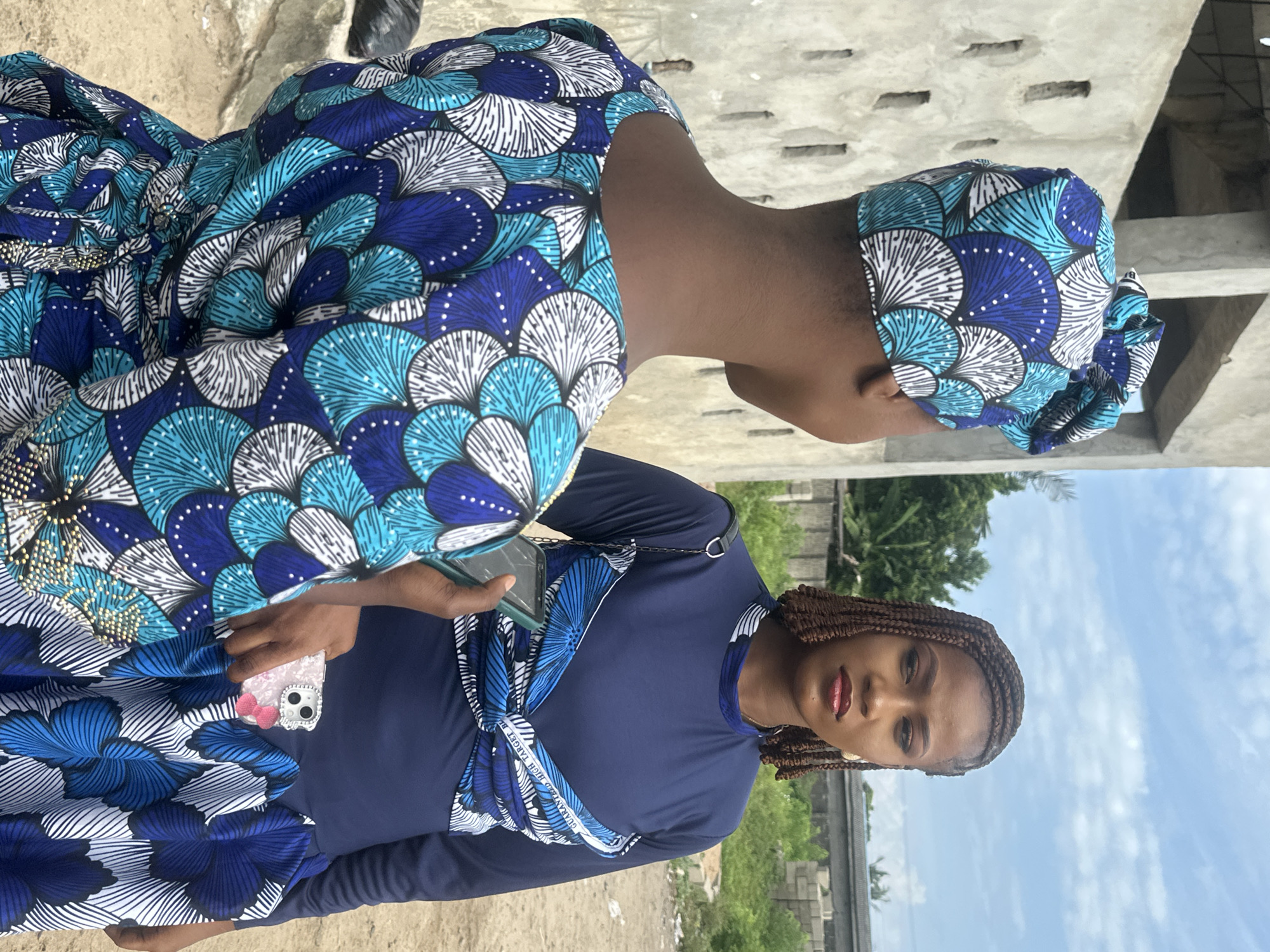 The image shows two women, one in the foreground and one partially visible in the background, in an outdoor setting that appears to be in Badagry, Nigeria. The woman in the foreground is wearing a dark blue long-sleeved dress with a prominent blue and white fan-like pattern on the chest area. She has braided hair and is holding a light pink phone case with a bow. Her expression is neutral. The woman in the background is wearing a similar patterned fabric as a headscarf and dress, also in shades of blue, white, and black. The setting suggests a daytime scene with a cloudy sky visible in the upper right portion of the image. The background includes what looks like unfinished concrete structures and some greenery, possibly indicating a construction site or a developing area. The ground appears to be dusty or sandy. There is some visible text on the blue fabric worn by the woman in the foreground, which reads "GUARANTEED HIGH TARGET" and "BEST". The overall impression is a casual outdoor moment.