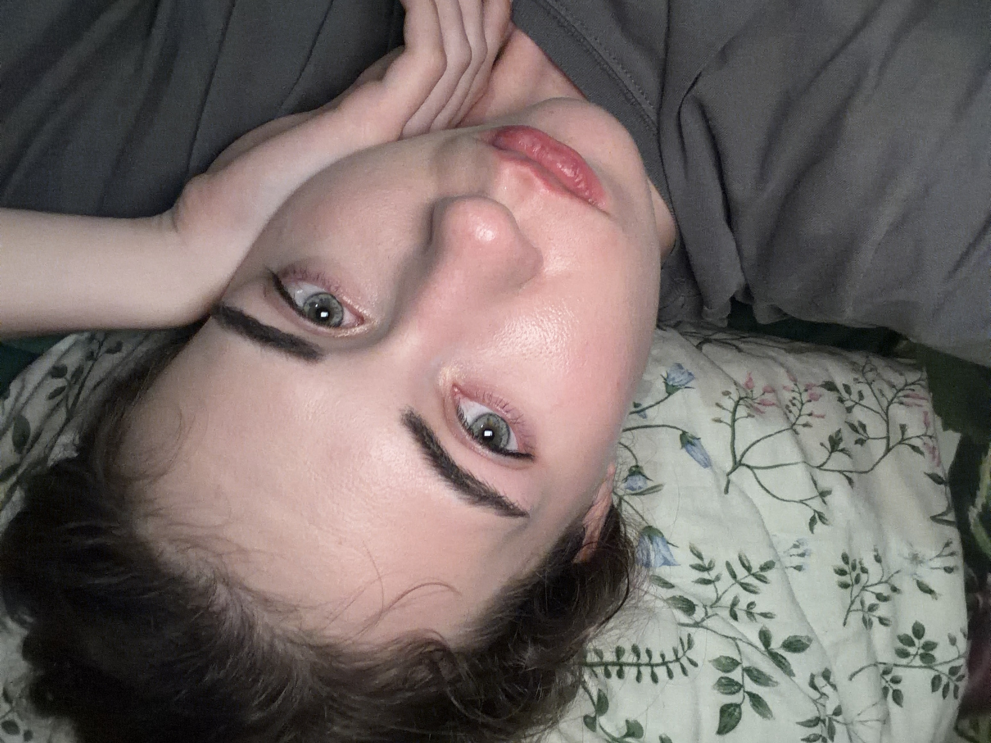 The image is a close-up, overhead shot of a person lying down. The person has fair skin, dark eyebrows, and light-colored eyes. Their head is tilted, and their left arm is bent, with a hand resting on their cheek. They are wearing a dark gray t-shirt. The background includes a floral-patterned fabric, possibly a pillow or blanket, with green leaves and blue and pink flowers. The scene appears to be indoors, with the lighting suggesting it is either late at night or in a dimly lit room. There is no discernible information about the city of Zavodoukovsk, Russia, in this image.