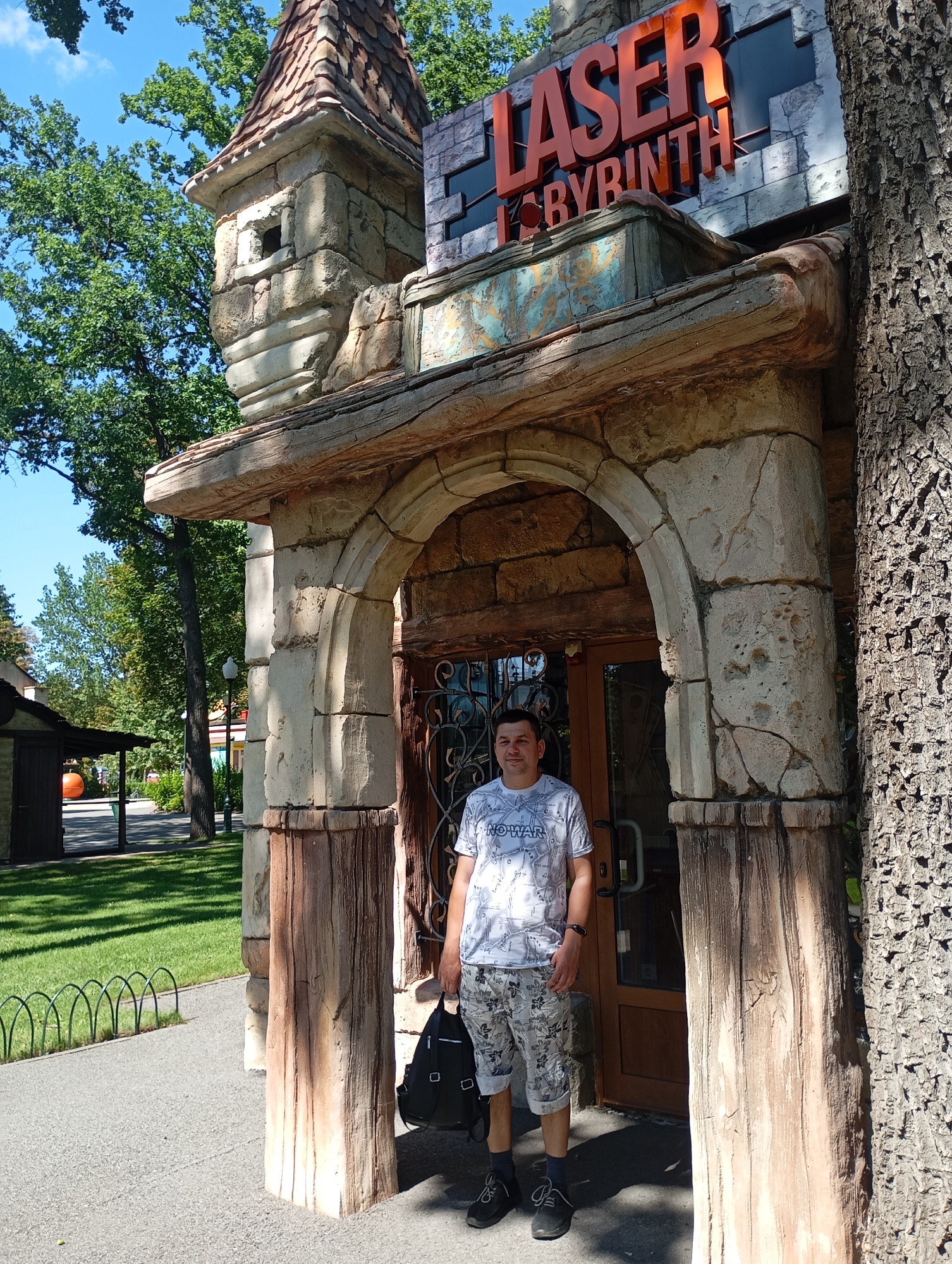 A man is standing in front of an arched entrance of a building that reads "LASER LABYRINTH" in orange letters. The building's facade is made of stone and faux wood, resembling a castle. The man is wearing a white t-shirt with a map-like pattern and the words "NO WAR," patterned shorts, dark socks, and black shoes. He is carrying a black backpack. The entrance is decorated with wrought iron. The scene is outdoors, with green trees and a blue sky visible in the background. The paved area in front of the entrance is a pathway. The overall atmosphere suggests a park or entertainment venue. The image appears to be taken in Kharkiv, Ukraine, as indicated by the "NO WAR" text on the man's shirt, a common sentiment expressed in Ukraine following the Russian invasion.