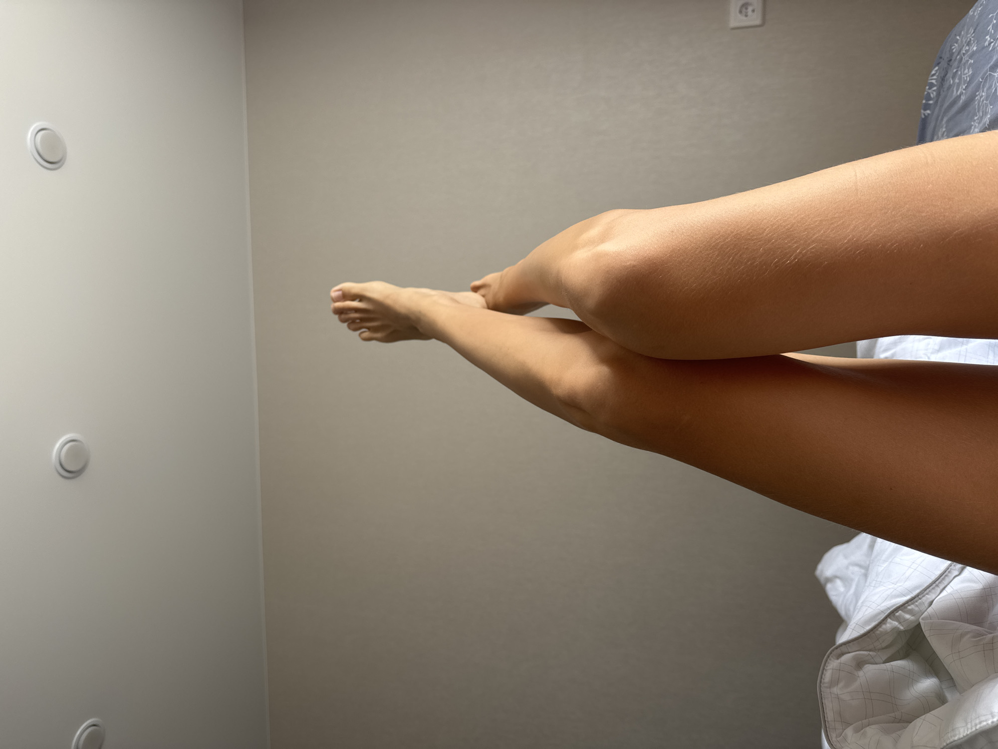 The image features a close-up view of a person's bare legs and feet, positioned diagonally across the frame, suggesting a relaxed posture such as resting on a bed or couch. The skin tone is tanned, and fine body hair is visible on the legs.
The setting appears to be an indoor room with neutral-colored walls, likely light gray or beige. On the left side of the frame, two circular, light-colored fixtures, possibly vents or lighting controls, are mounted vertically on the wall. A standard electrical outlet is visible on the wall in the upper right quadrant. Part of a white duvet or blanket with a subtle checkered or grid pattern is visible on the lower right, suggesting a bed.
The lighting is bright and even, casting minimal shadows, which could indicate natural daylight or well-distributed artificial light. No specific activity is taking place beyond the person resting their legs. There are no discernible emotional cues or visible text within the image.