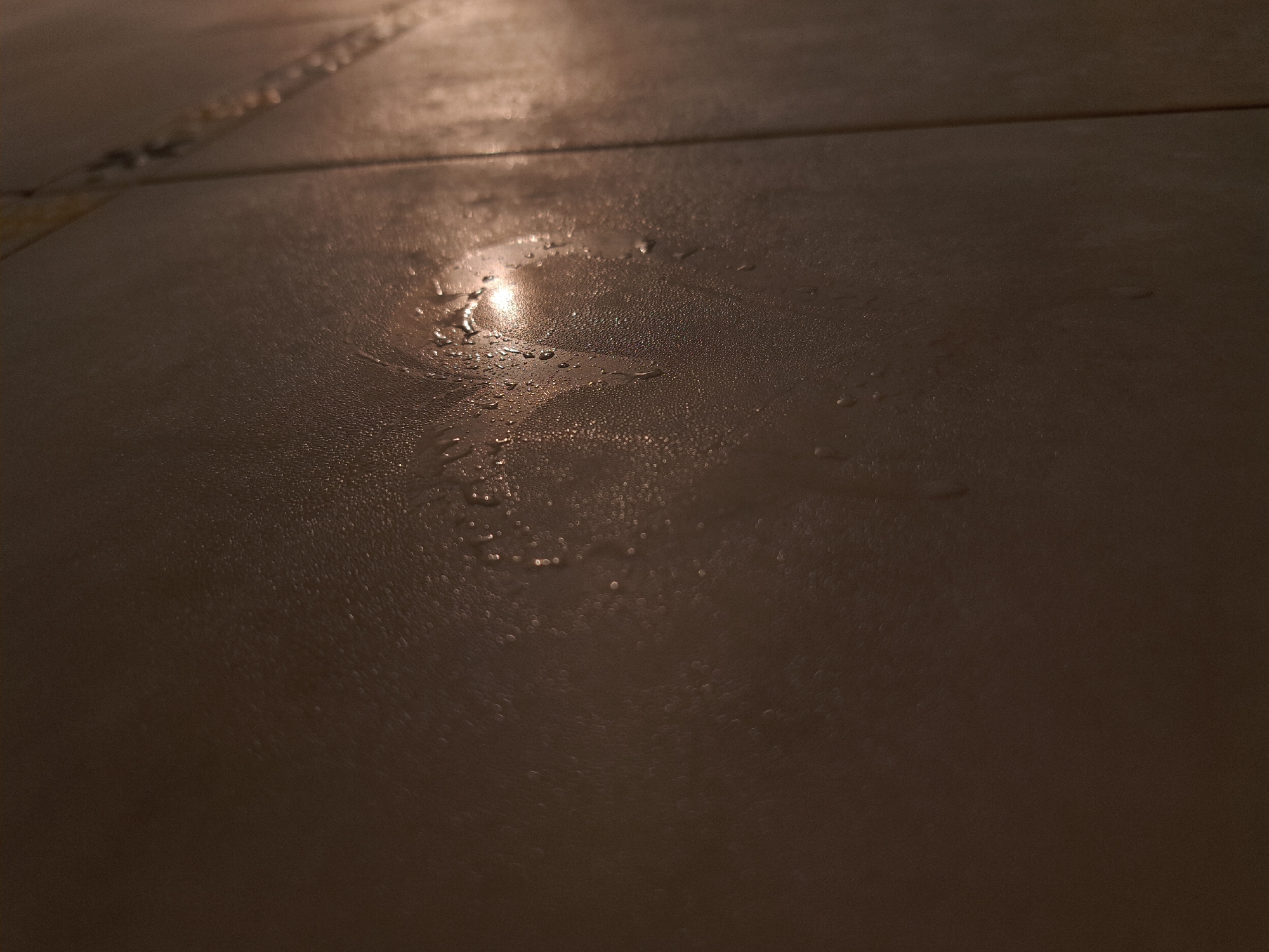The image shows a close-up of a section of tiled floor, likely indoors. The tiles are brown and slightly reflective. There are water droplets or condensation scattered across the surface of the tiles. There are no people, subjects, or other objects visible. The environment is dark. There is no information available to determine if the image is located in Negovan, Bulgaria.