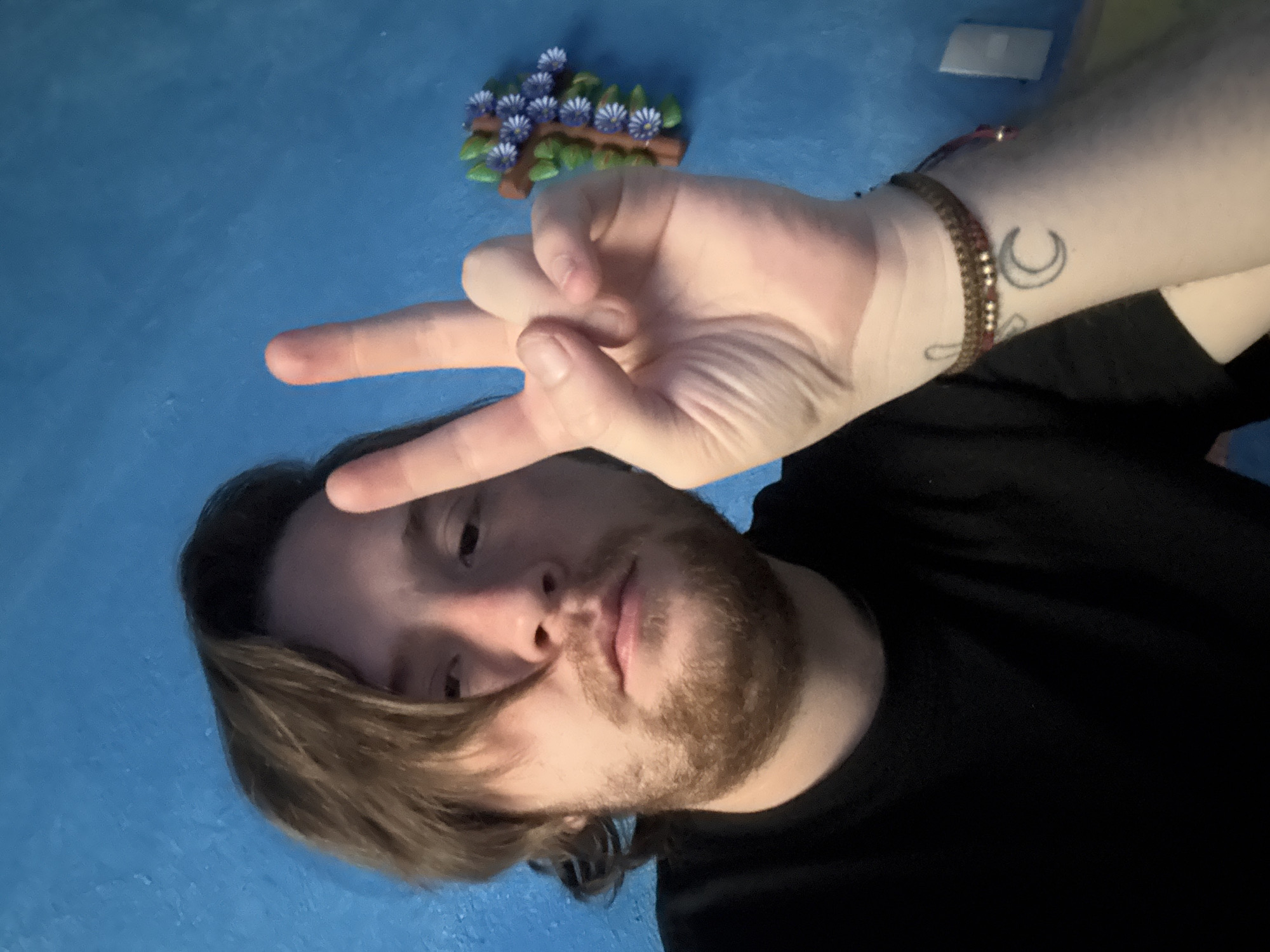 A man with light brown hair and a beard is making a peace sign with his right hand in front of a blue background. He is wearing a black shirt. A small decorative object with purple flowers is in the upper left corner of the image. A bracelet and crescent moon tattoo are visible on the man's right wrist.
The location is not discernible from the image.