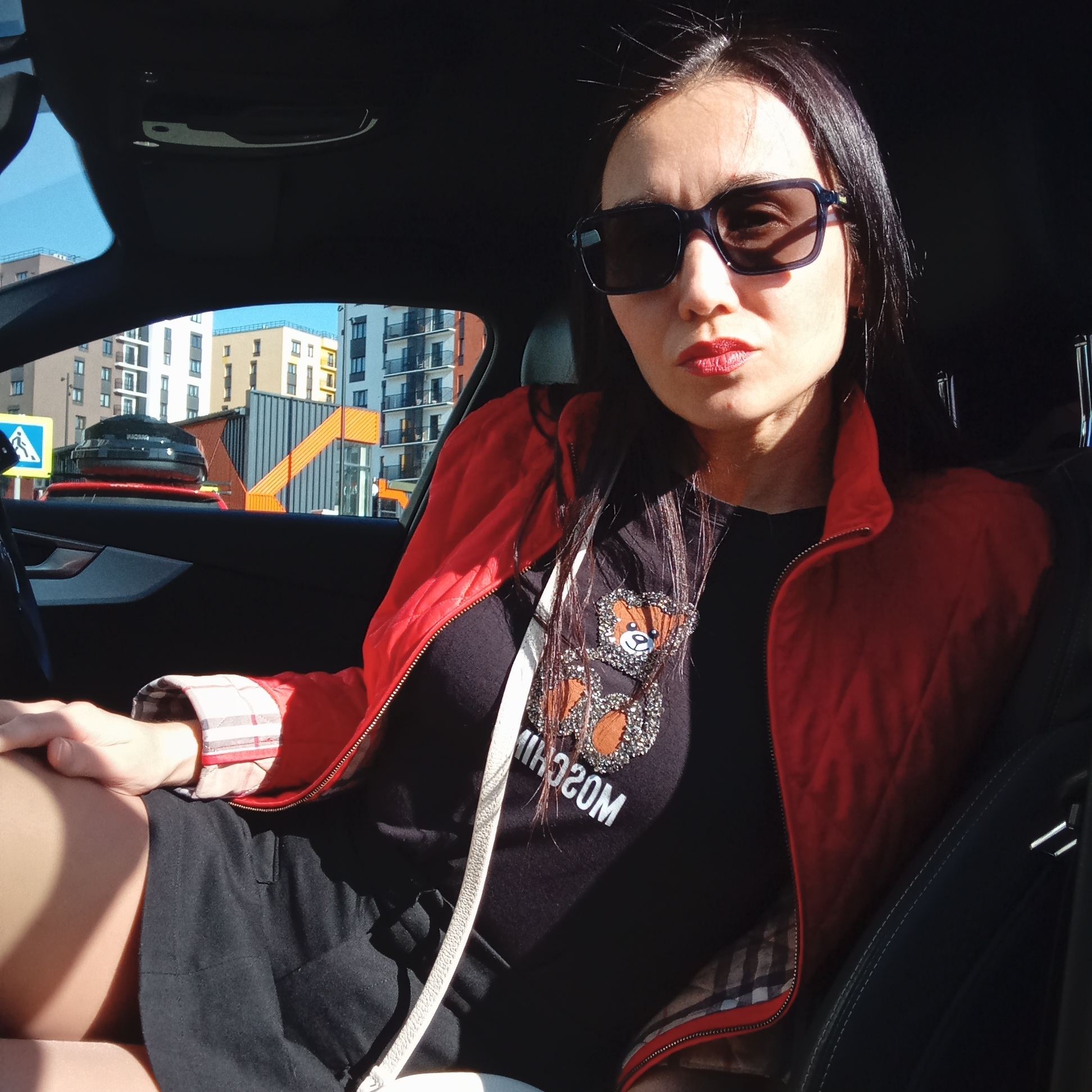 A woman with dark hair, wearing sunglasses, a red jacket, and a black t-shirt with a cartoon bear design, is sitting inside a car. The car is parked in an urban environment in Pervomaysky, Russia. Multi-story apartment buildings and a pedestrian crossing sign can be seen through the car window.