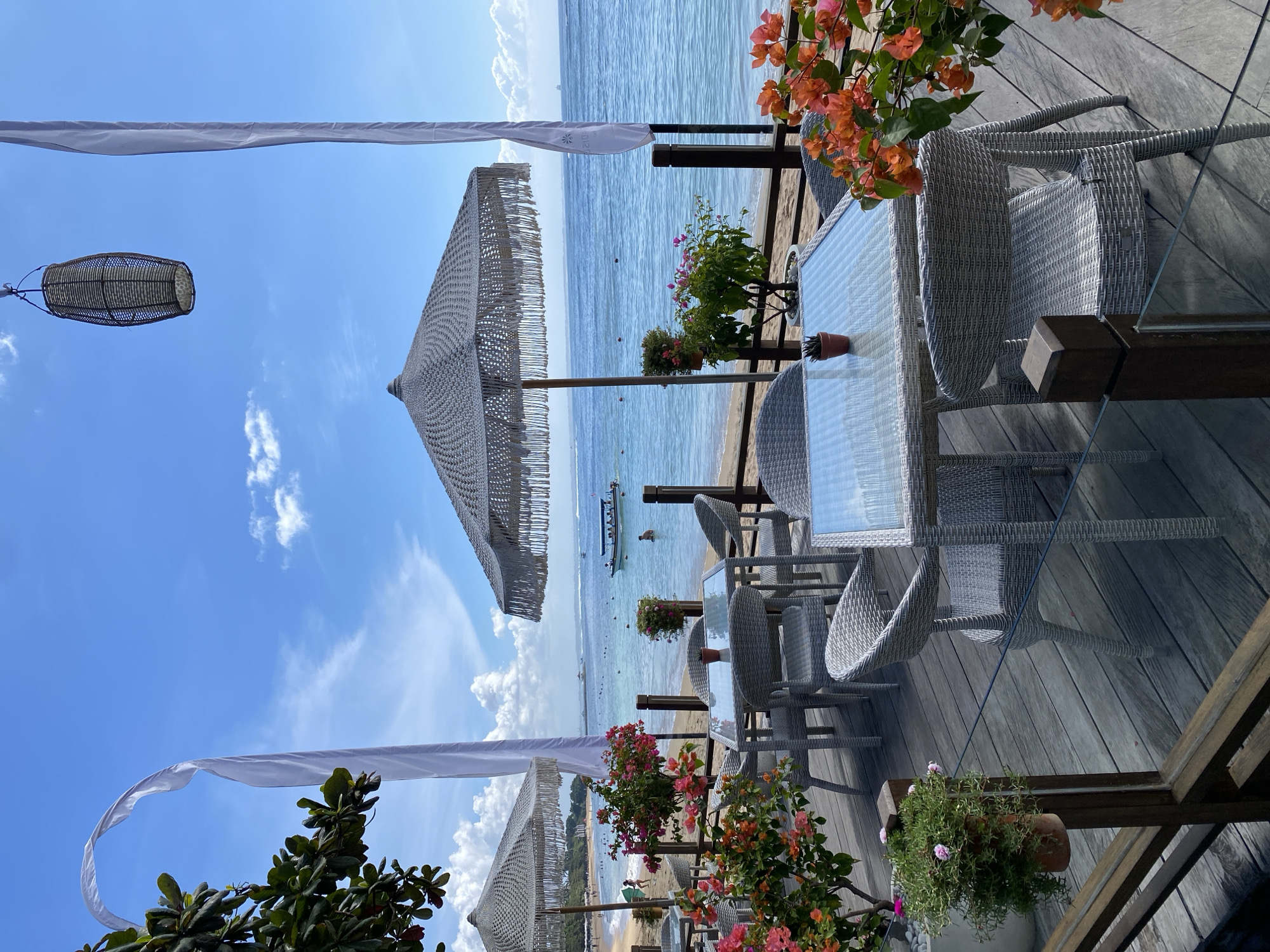 The image shows a seaside restaurant in Kuta Selatan, Indonesia. The foreground contains a wooden deck with white wicker furniture arranged around glass-topped tables. Pots of colorful flowers, including bougainvillea, decorate the tables and deck edges. Woven sun umbrellas and white fabric draping provide shade. Beyond the deck is a sandy beach and the ocean, with boats visible in the distance. The sky is blue with scattered clouds.