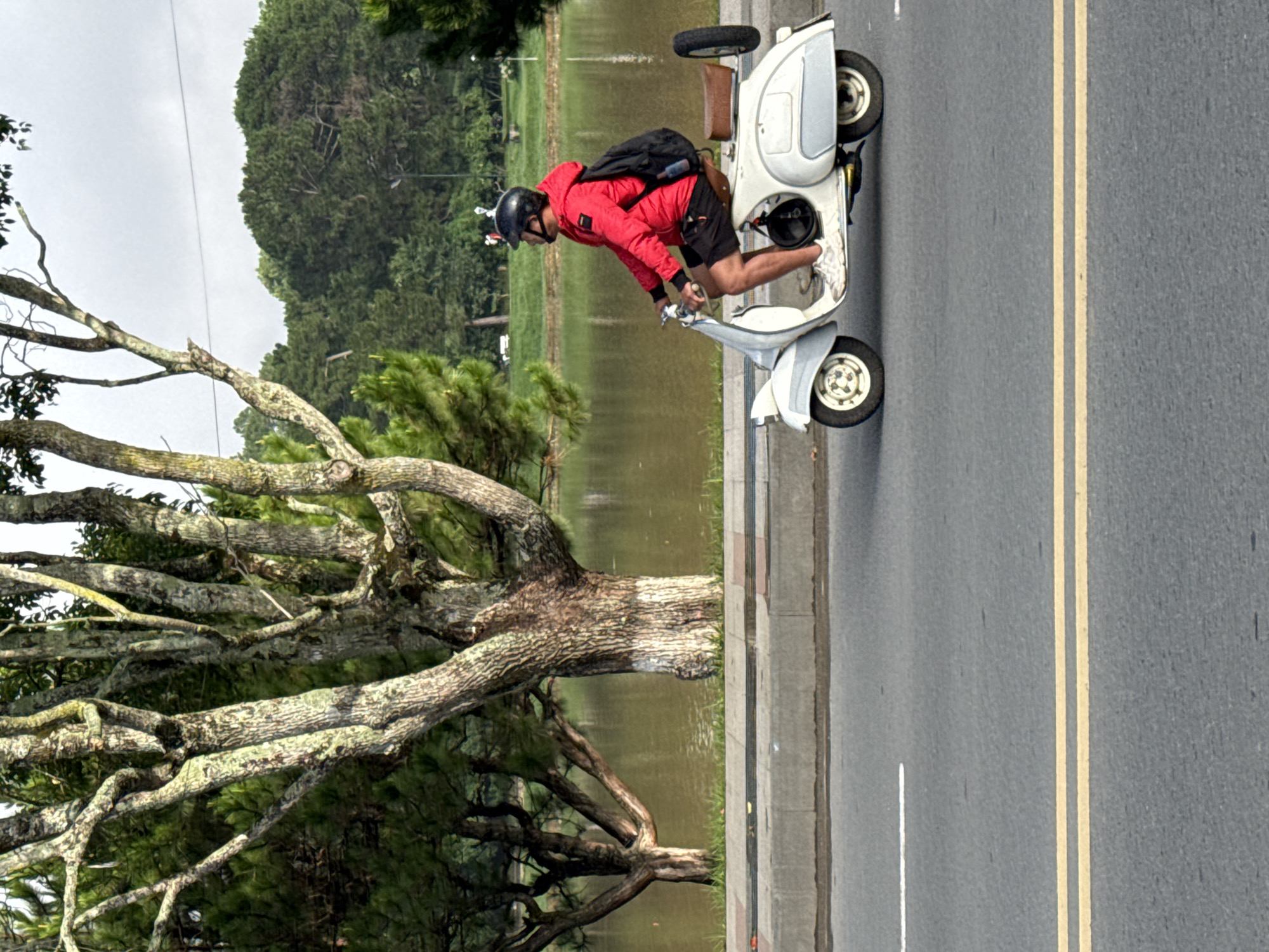 A man wearing a red jacket, black helmet, and black shorts is riding a white scooter on a paved road. He has a black backpack on and is looking to his left. In the background, there is a body of water, trees, and a cloudy sky. The scooter has a spare tire mounted on the back. The scene appears to be in Da Lat, Vietnam, likely near Xuan Huong Lake, given the presence of the lake and surrounding pine trees, which are characteristic of the city's landscape.