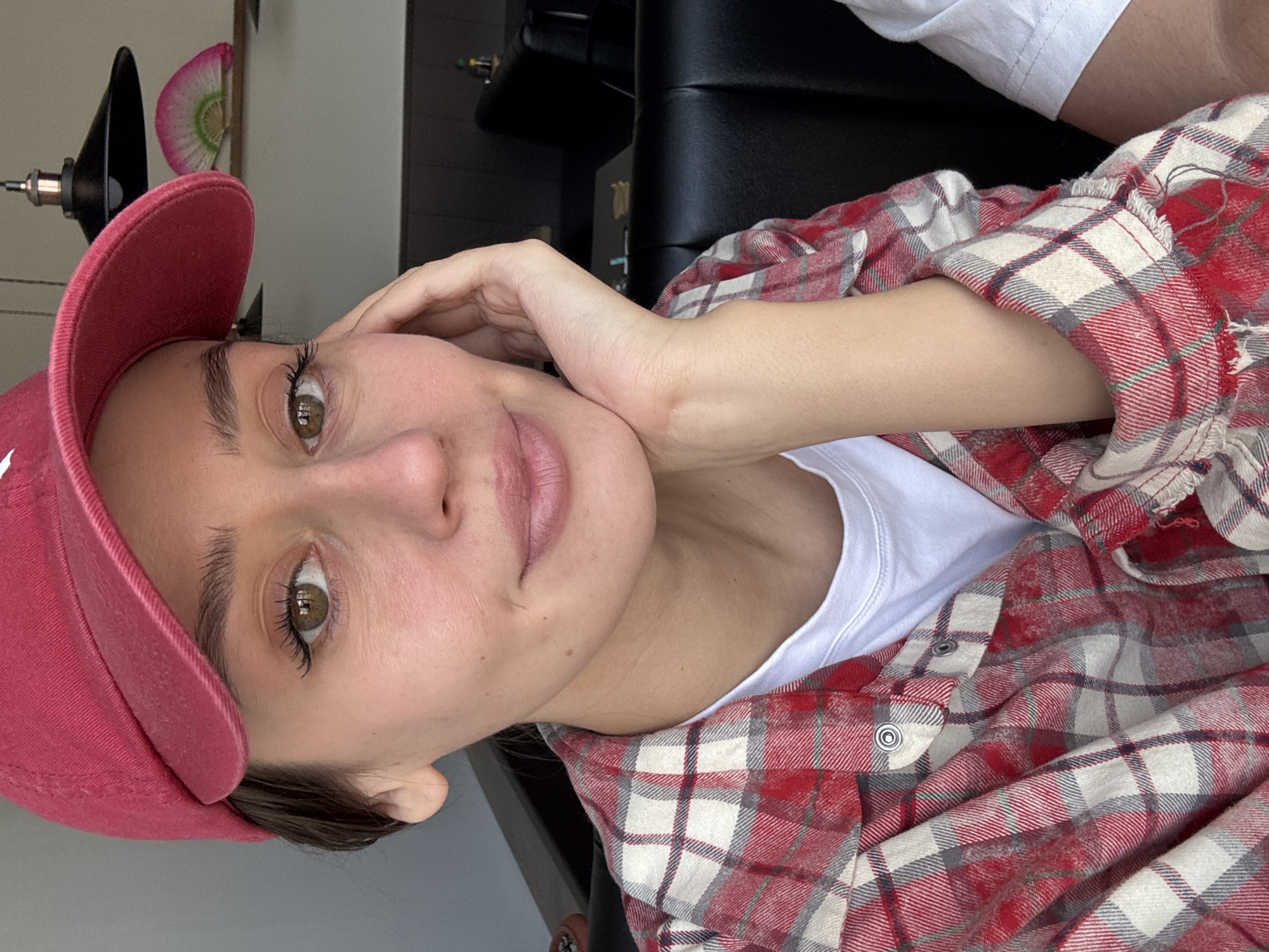 A close-up portrait shows a person, likely a young woman, taking a selfie in an indoor setting. The image is oriented with the subject's head to the left, looking directly into the camera with a neutral, relaxed expression.
The person is wearing a bright red baseball cap, which partially covers their dark hair. Their eyes are a striking light brown or hazel, accentuated by long, dark eyelashes and well-defined eyebrows. Their lips have a natural, soft pink coloration. One hand is gently resting against their cheek and jawline.
For attire, the person is wearing a red, white, and grey plaid flannel shirt, worn open over a plain white crew-neck t-shirt. The texture of the flannel fabric is visible, including some frayed edges on the shirt.
The background is softly blurred but reveals elements of an indoor space. To the left, a dark metallic fixture, possibly part of a lamp or furniture, is visible. Behind the subject's head, a small, decorative, fan-shaped object with pink and green patterns can be seen. Further in the background, dark, possibly leather, furniture or upholstery is discernible. The lighting appears soft and natural, suggesting daylight. No visible text is identifiable on the clothing or in the background.