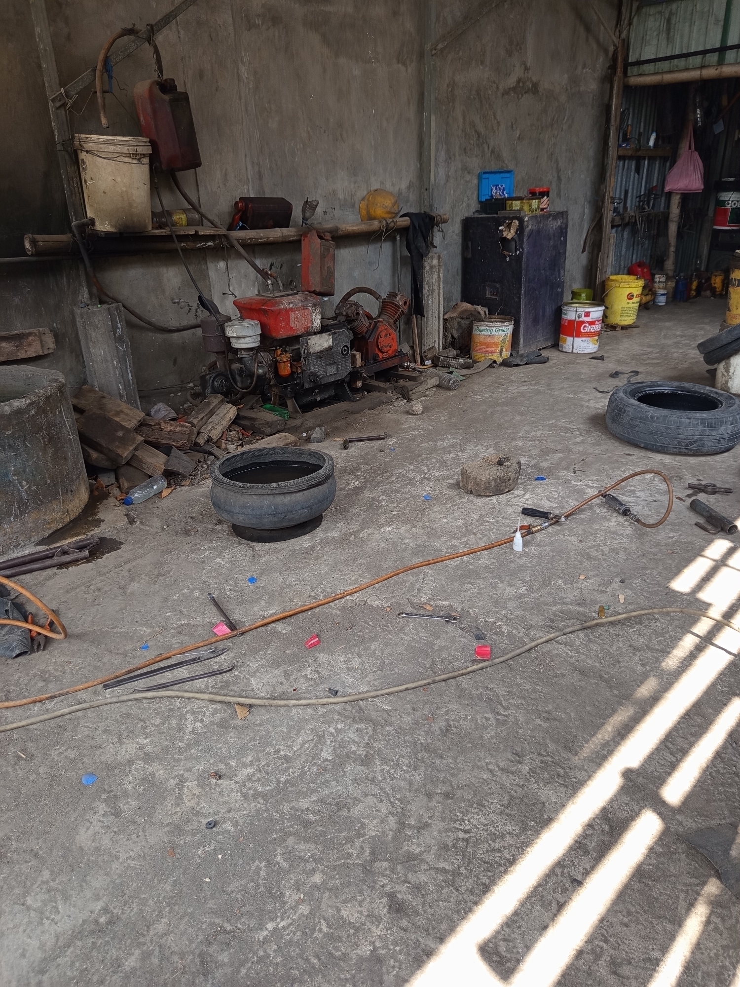 This is a photograph of the interior of a workshop in Mrican, Indonesia. The workshop is a cluttered space with a concrete floor and rough concrete walls. In the center of the image, a small red engine is visible. Various tools and equipment are scattered around the floor, including tires, hoses, and cans. There are also several plastic bottles and pieces of debris. In the background, a shelf holds various containers, including what appear to be fuel cans and a white bucket. The lighting suggests it is daytime, with sunlight casting strong shadows across the floor from a window or opening. The overall impression is of a working garage or maintenance area that is in active use and has not been recently cleaned.