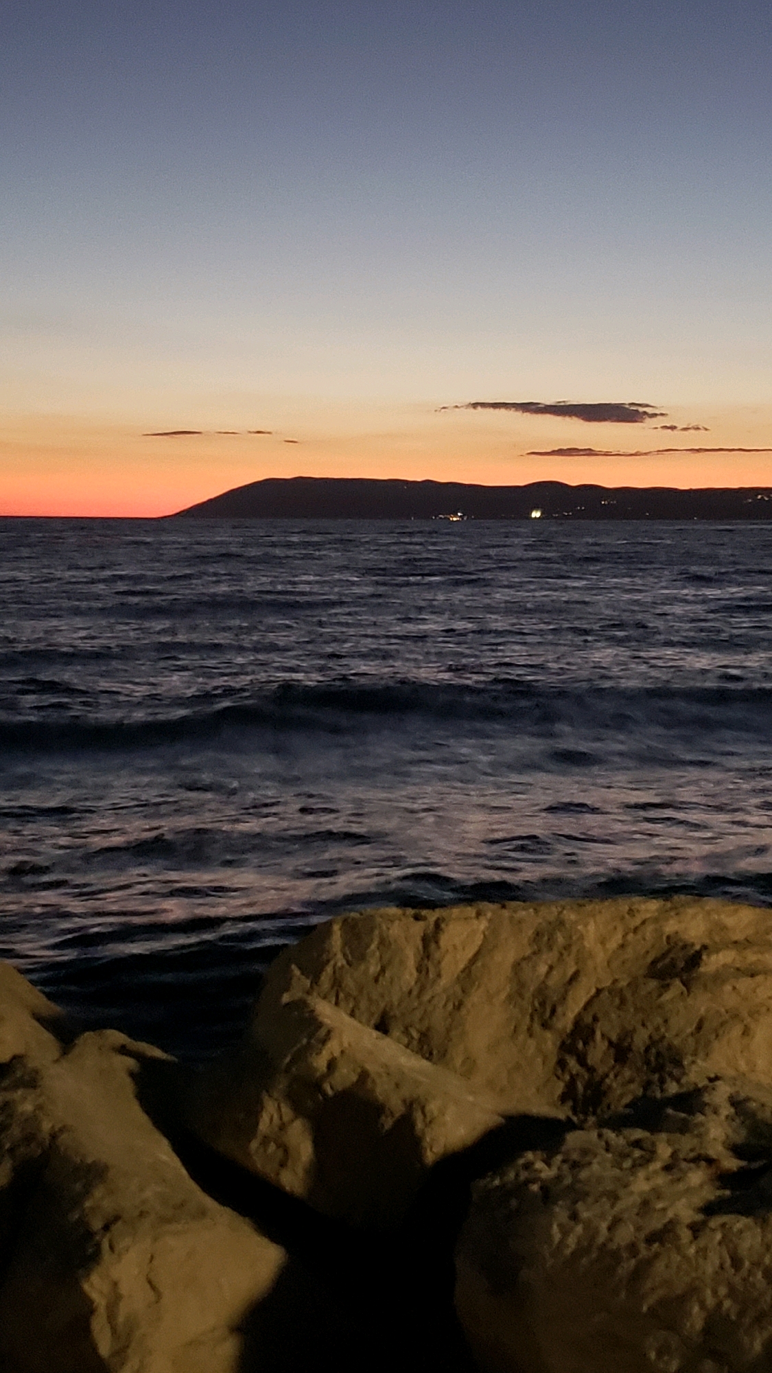 The image depicts a twilight scene at the coast of Općina Tučepi, Croatia. In the foreground, large, light-colored rocks are visible, illuminated from the side, casting deep shadows. Beyond the rocks, the dark, rippling surface of the sea occupies the middle ground, with visible waves creating a sense of movement. In the distance, a dark silhouette of a landmass, likely a mountain or island, is seen against the horizon. The sky above transitions from a soft orange and pink glow near the horizon, indicative of a sunset or sunrise, to a pale blue at the top of the frame. A few wispy clouds are scattered across the sky, and faint lights are visible on the distant landmass, suggesting human settlement. The overall atmosphere is calm and serene, characterized by the natural elements of water, rock, and sky.