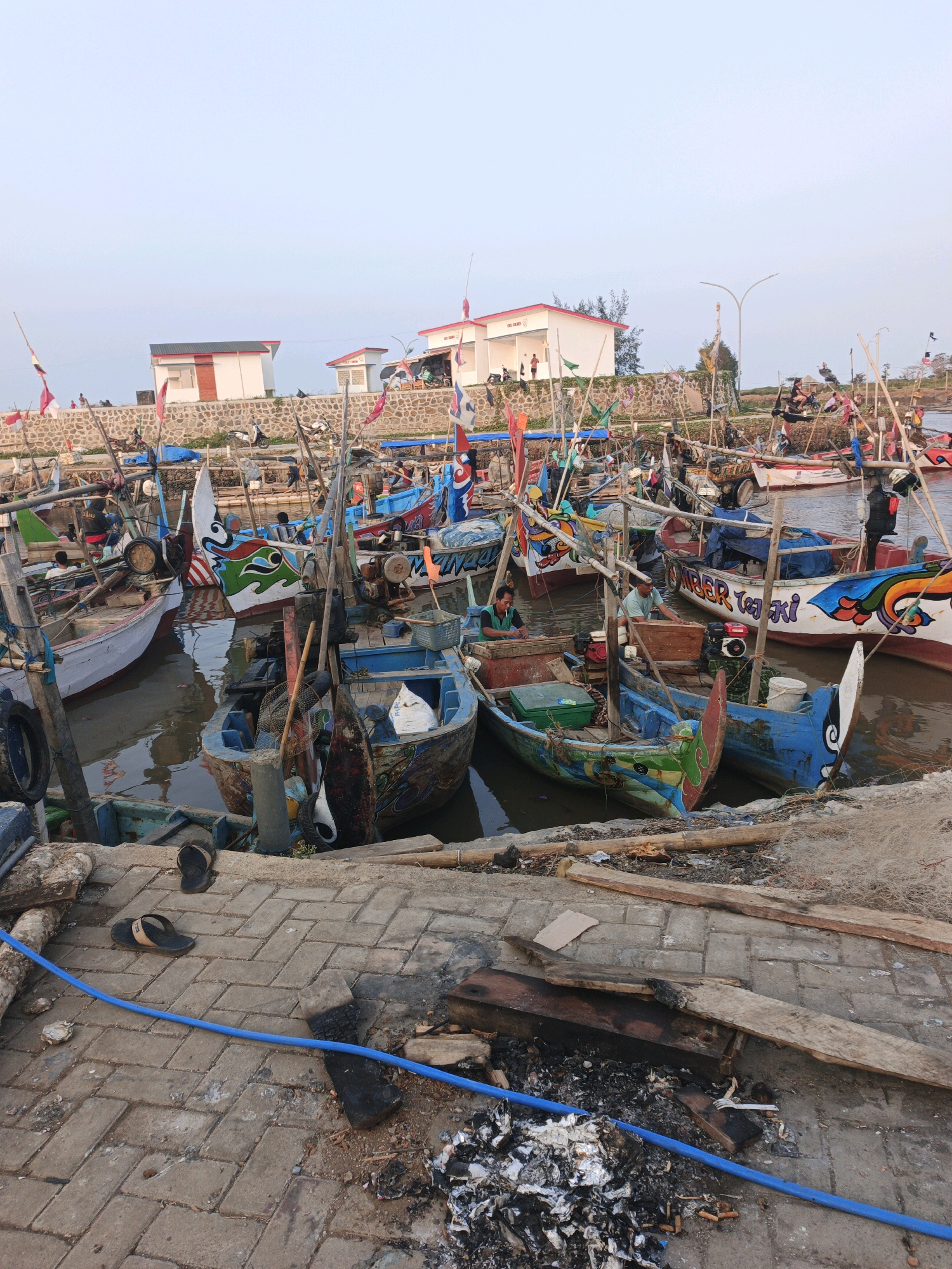 The image shows a harbor scene in Bumiharjo, Indonesia, filled with numerous small fishing boats. The boats are docked closely together in the water and are brightly decorated with various colors and designs. Several people are visible on and around the boats, some appearing to be preparing for or returning from fishing. The foreground shows a paved area with debris, including discarded wood, a blue hose, and a pair of flip-flops. In the background, there are buildings with red roofs and a stone wall, suggesting a coastal settlement. The sky is clear and a light blue, indicating daytime. The overall impression is one of a bustling fishing community.
