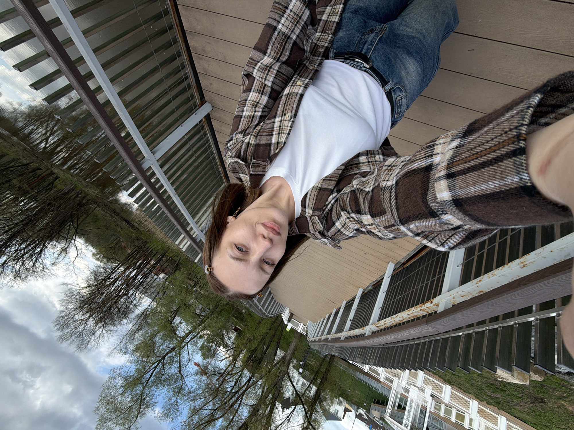 A woman is taking a selfie outdoors. She is wearing a white t-shirt, a brown and tan plaid flannel shirt, and blue jeans. A black belt is visible at her waist. The setting appears to be a wooden walkway or bridge, with metal and wood railings. Trees and building structures are visible in the background. The photo's orientation is slightly tilted. According to geolocation, the location is in Klin, Russia.