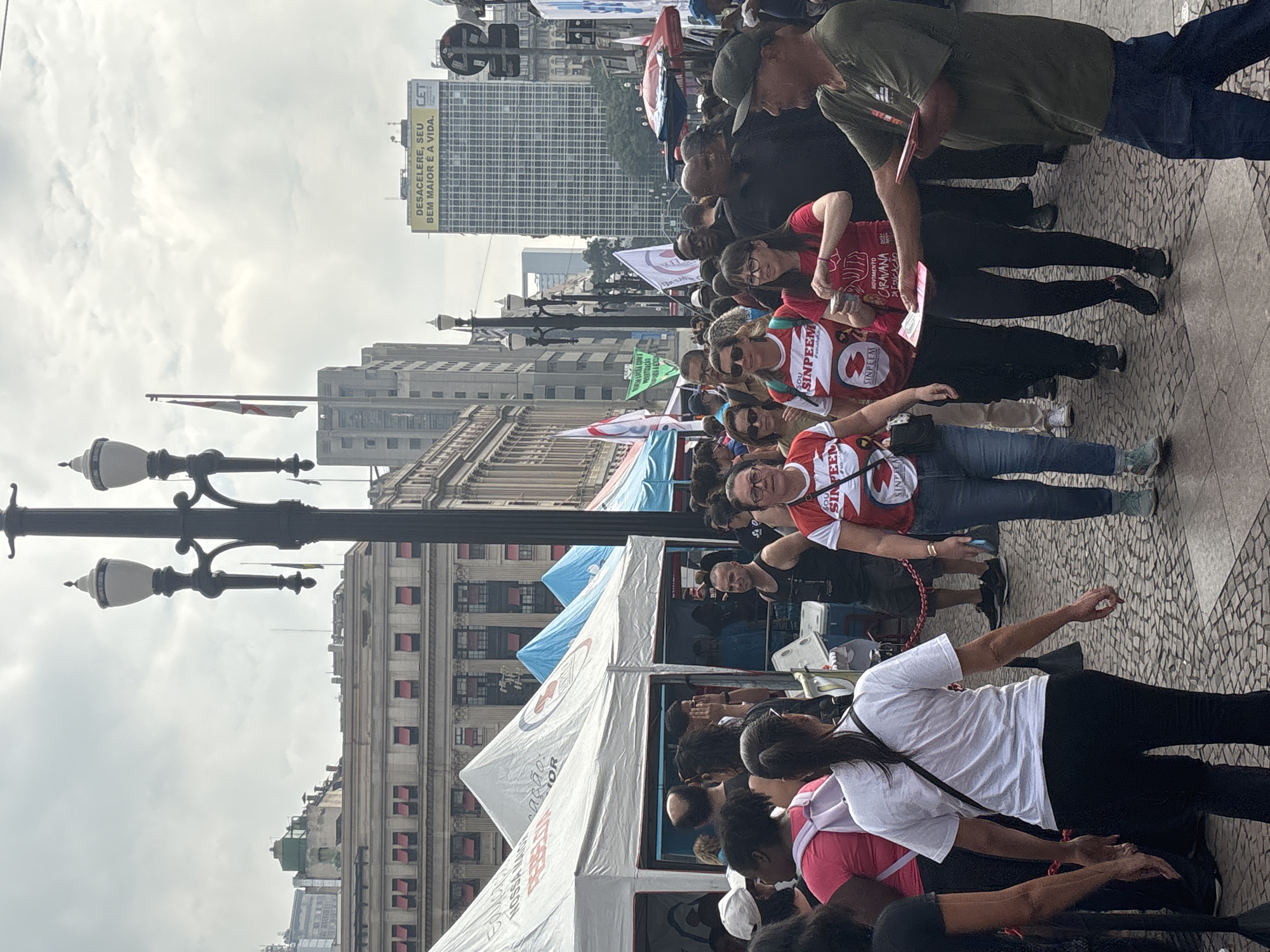 This vertical image captures an outdoor gathering or event in an urban environment in São Paulo, Brazil. The scene depicts numerous people, mostly adults, assembled on a paved street or public square, with tall city buildings and ornate street lamps forming the backdrop under an overcast sky during the daytime.
Many participants are wearing matching red and white shirts or vests with "SINPEEM" and "SOU SINPEEM" visible on them, suggesting an organized group, likely an educational workers' union. Some individuals are seen looking at papers or interacting within their groups. In the mid-ground, white and blue event tents are set up, contributing to the sense of a planned public assembly. A flag or banner partially displays "MOVIMENTO CARAVANA EDUCAÇÃO" (Education Caravan Movement), reinforcing the theme of the gathering.
A prominent billboard on a distant skyscraper features the Portuguese text: "DESACELERE, SEU BEM MAIOR É A VIDA. CET," which translates to "Slow down, your greatest good is life. CET." The overall mood appears orderly and focused, with participants engaged in the event's activities.