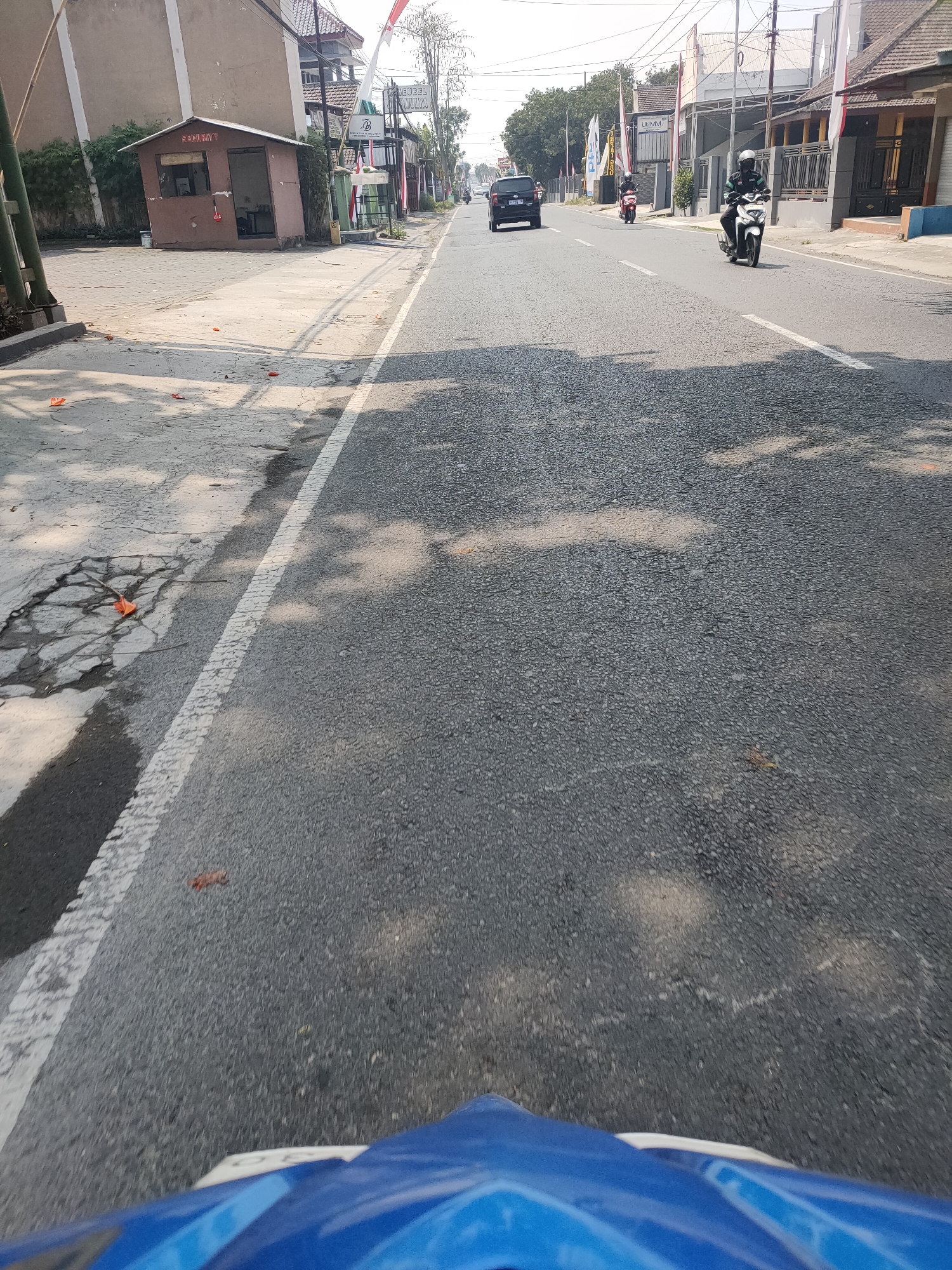 The image captures a street scene in Kanigoro, Indonesia, likely from the perspective of someone riding a motorcycle. The road is asphalt, with a white dashed line marking the lane. To the left, a gray concrete sidewalk is visible, featuring cracked sections and some fallen orange leaves. Adjacent to the sidewalk is a small, brown security booth with "SECURITY" signage. Further down the road on the left, several buildings and trees line the street. On the right side of the road, houses and shops are visible. Traffic includes a black SUV in the distance and two motorcycles in motion, one white and one red, with riders wearing helmets. The sky is bright and clear, indicating daytime. The foreground shows a portion of a blue motorcycle fairing, suggesting the photo was taken from a moving vehicle.