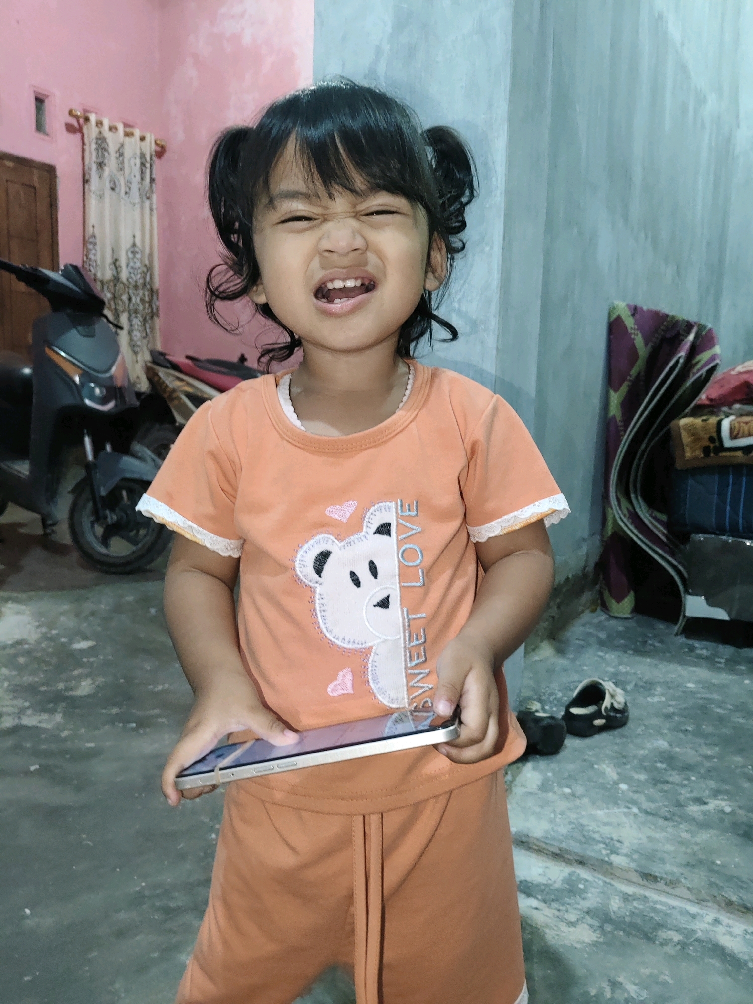 In Fajar Baru, Indonesia, a young Indonesian girl with dark hair styled in pigtails grimaces while holding a smartphone. She wears an orange t-shirt with a teddy bear design and the text "SWEET LOVE," paired with matching orange shorts. The background reveals a pink wall with a wooden door and curtains on the left, and a textured grey wall on the right. A scooter is parked to the left of the girl. The floor is a mottled concrete surface.