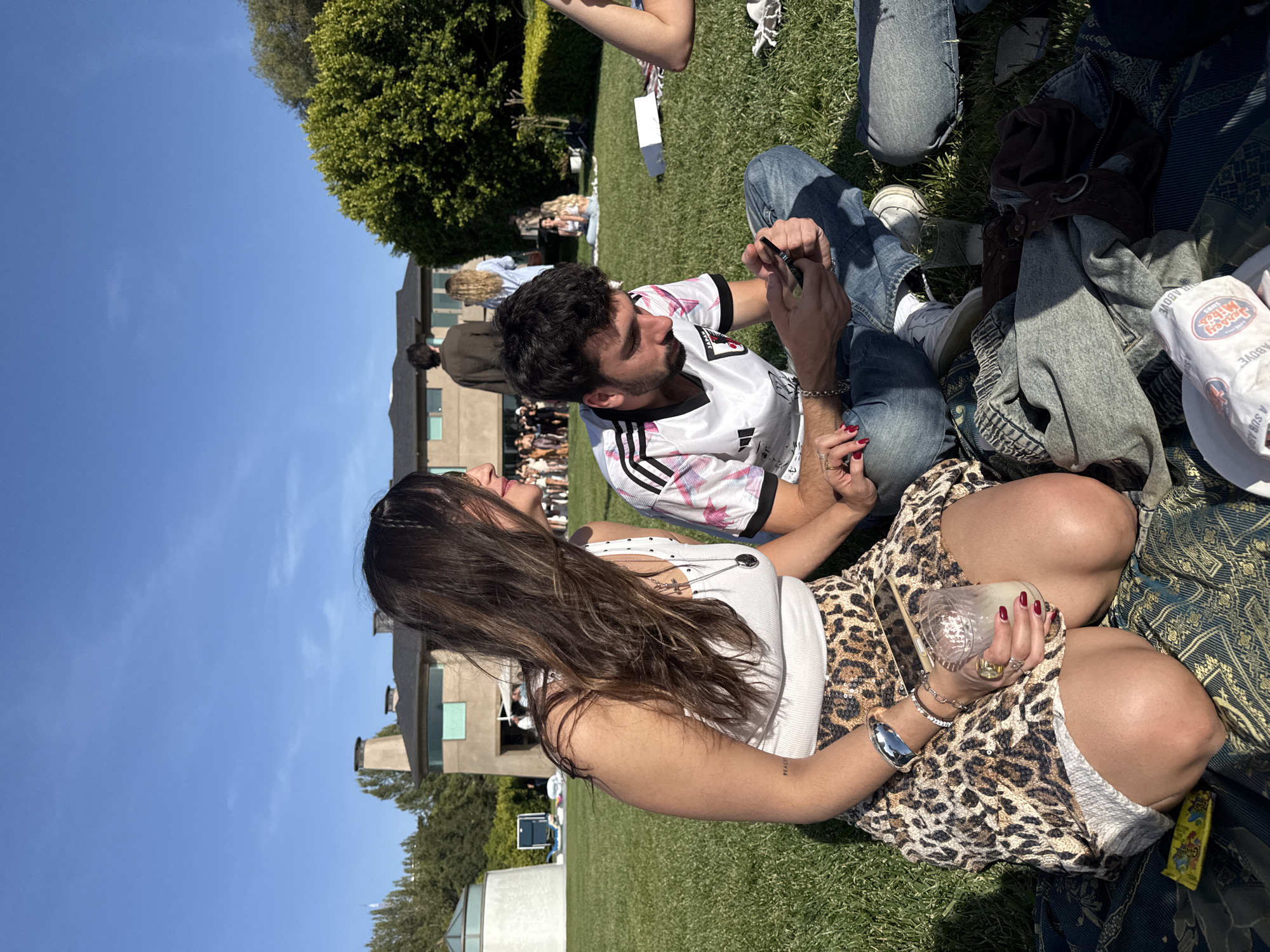 The image captures an outdoor scene on a sunny day in Los Angeles. Several people are seated on a grassy lawn. A man wearing a white soccer jersey with black stripes and a pink floral pattern is focused on his smartphone. He is seated next to a woman wearing a white tank top and a leopard-print skirt, holding a glass. They seem to be engaged in a close interaction. Other individuals are scattered around them, some seated on blankets and others standing in the background. A large building is visible in the background.