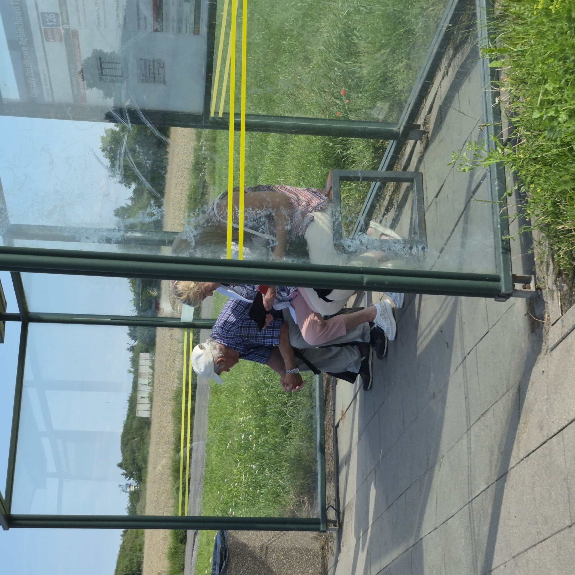 The image shows a bus stop shelter with three people sitting on a bench. The shelter has green metal framing and glass panels. Inside the shelter, from left to right, a man wearing a blue checkered shirt, grey pants, and a white cap is seated. He is holding a dark object in his hands. Next to him, a woman wearing a pink and white patterned top and white pants is seated. To her right, another woman, largely obscured by a smudge on the glass, is also seated. The bus stop shelter is situated outdoors, with a grassy area and a field visible behind it. The ground in front of the shelter is paved with grey concrete slabs. A dark grey trash bin is visible to the left of the shelter. A sign is visible on the interior of the right-hand glass panel, showing route information, including "126". This bus stop is located at "Ogordy Dzialkowe Rebiechowo I" in the city of Gdańsk, Poland. The overall scene depicts a sunny day at a public transportation stop.