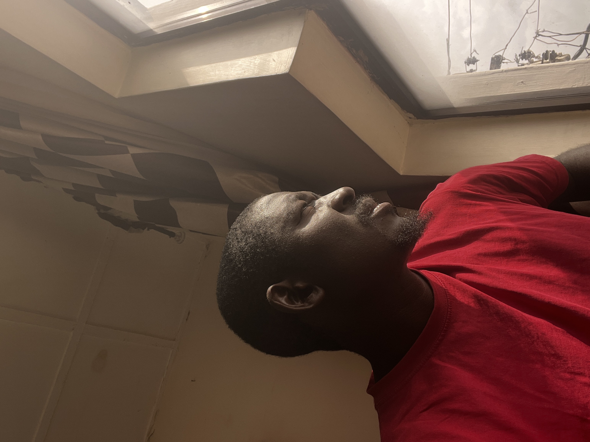 A close-up, side profile shot features a Black man wearing a red t-shirt, his head turned to the right, looking out a window. His eyes are partially closed, and he has a goatee. The room's walls appear to be a muted yellowish-brown, and the ceiling is visible in the upper left corner. A checkered flag pattern is present in the upper right portion of the image, hanging loosely. The window frame is light brown and appears to be made of wood. Outside the window, electrical wires and poles are visible, suggesting an urban or semi-urban setting. The lighting indicates it is daytime, possibly overcast. The image is likely taken in Jos, Nigeria, based on the context provided.
