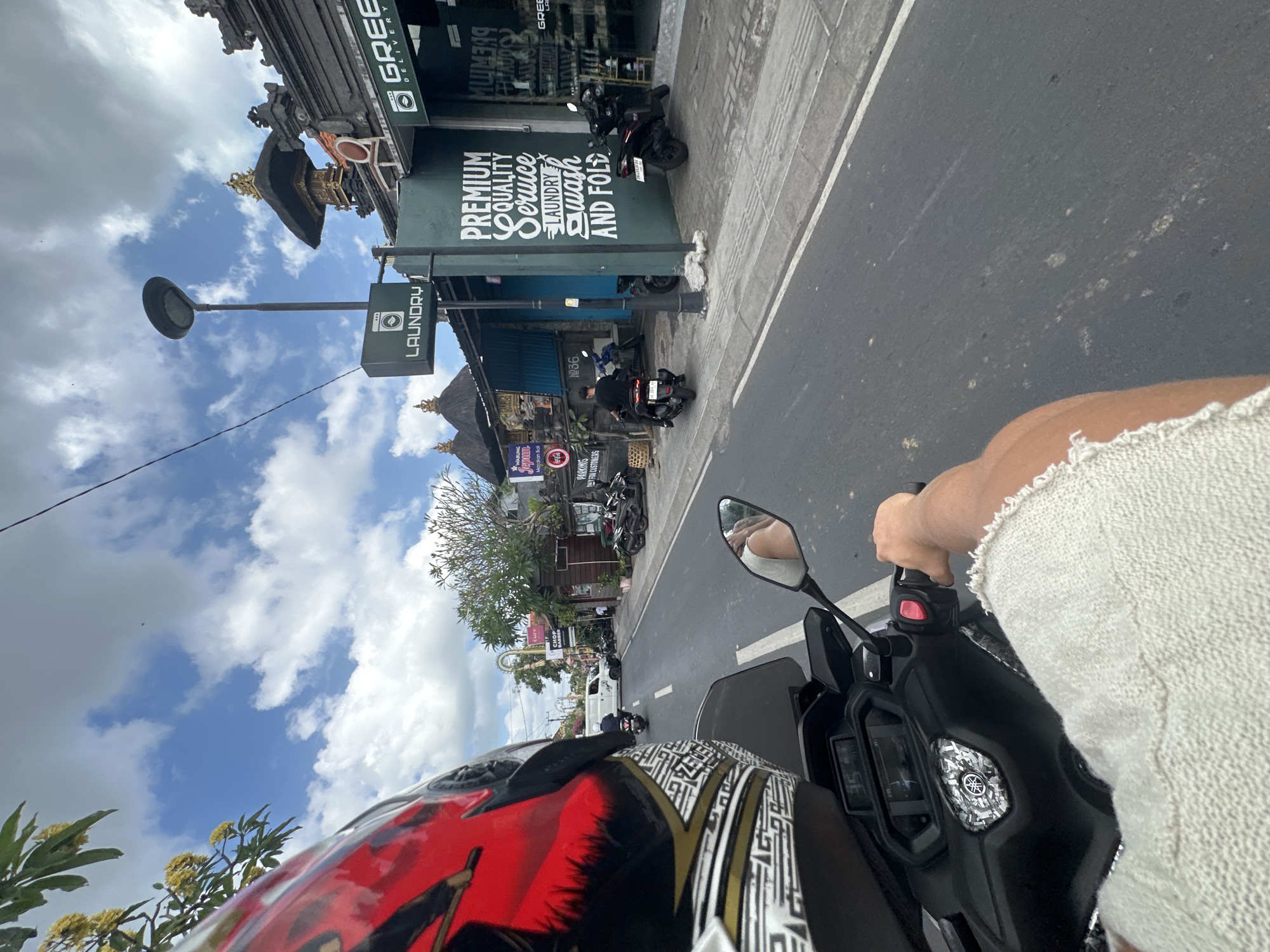 The image is a first-person perspective of someone riding a scooter on a street in North Kuta, Indonesia. The rider is wearing a red and black helmet and a light-colored, textured shirt. In the foreground, the handlebars and dashboard of the scooter are visible, showing a digital display with the number "35". The road is paved and has a white dividing line.
On the right side of the street, there is a green building with signage that reads "PREMIUM QUALITY Service LAUNDRY WASH AND FOLD" and "GREEN DELIVERY". There are several parked motorcycles in front of this building. Further down the street, a white van is visible, and other buildings line the road.
On the left side of the street, there is a tree with yellow flowers. Above, the sky is blue with scattered white clouds. The environment appears to be a typical urban street scene with commercial establishments and traffic.