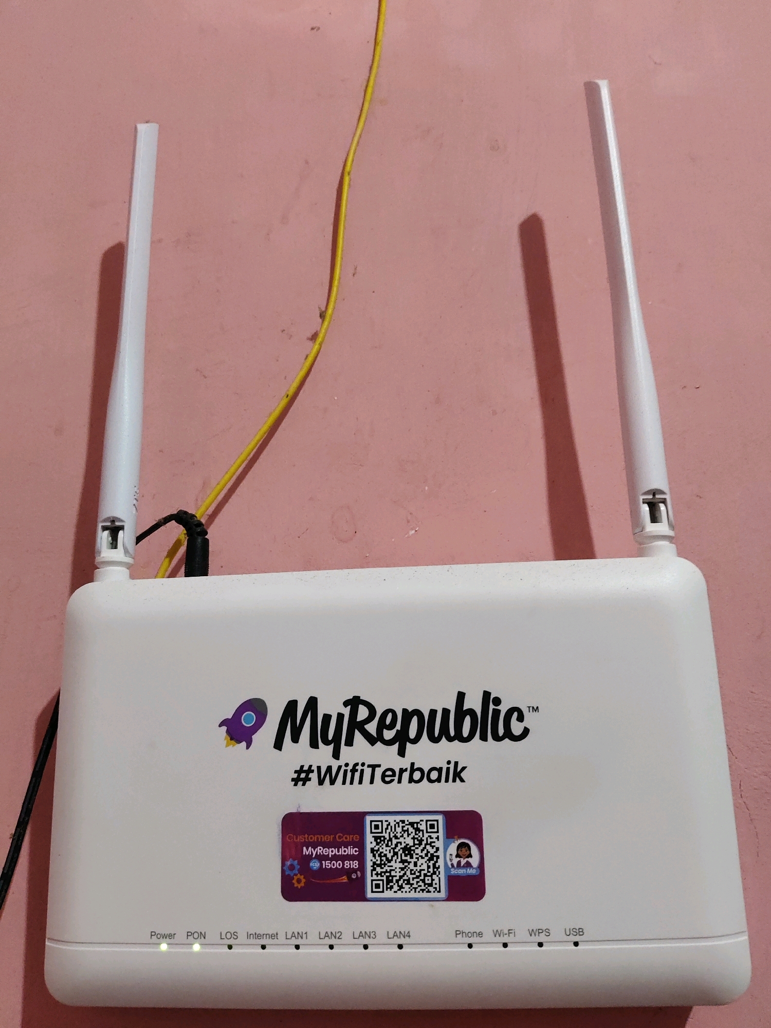 The image shows a white MyRepublic router with two antennas, placed against a light pink wall. A yellow cable is visible running along the wall behind the router, and a black cable is plugged into the router. There are several stickers on the front of the router, including a rocket logo, the text "MyRepublic", "#WifiTerbaik", a "Customer Care MyRepublic 1500 818" sticker, and a QR code with a "Scan Me" sticker. Below the stickers, there are various indicator lights and labels for "Power", "PON", "LOS", "Internet", "LAN1", "LAN2", "LAN3", "LAN4", "Phone", "Wi-Fi", "WPS", and "USB". The "Power" and "PON" lights are illuminated green. The router is positioned in an indoor setting. The location of Fajar Baru, Indonesia is not determinable from the image.