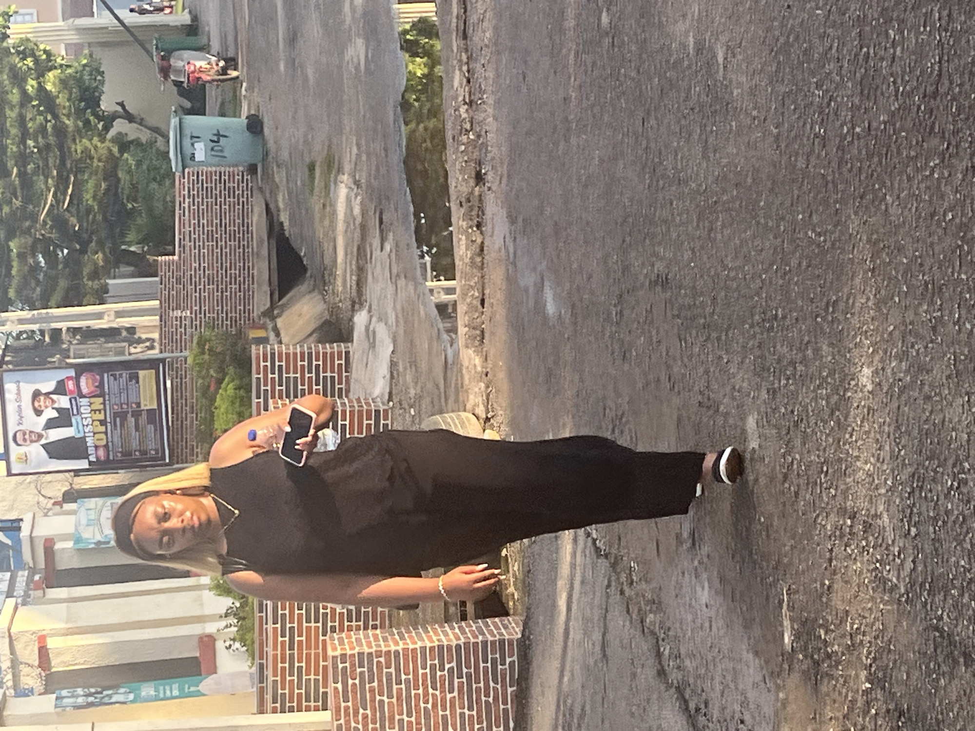 A Black woman with blonde-streaked hair, wearing a black sleeveless dress and a black headband, walks away from the camera on a paved street. She holds a water bottle and a smartphone in her right hand. Her left arm is slightly bent, and a bracelet is visible on her left wrist. She wears black and white shoes. The street is wet, with puddles reflecting the environment. To the left of the street, there is a brick wall and a building with columns. Further back, there is a green trash can with the number "104" spray-painted on it, a red motorcycle, and trees. The sky is visible in the upper right portion of the image. The lighting suggests it is late afternoon. The scene appears to be an outdoor urban environment in Abuja, Nigeria.
