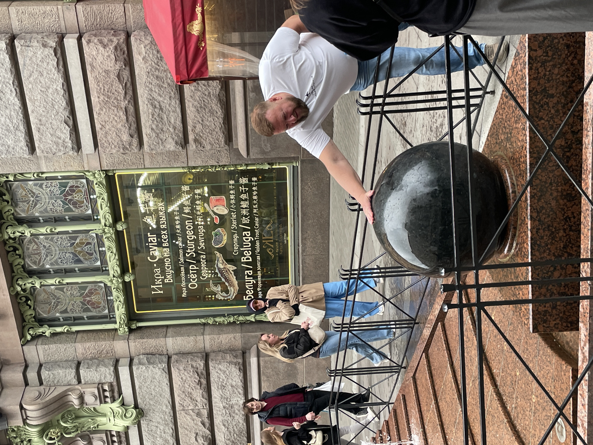 The image depicts a scene in Saint Petersburg, Russia. A man in a white shirt and jeans is interacting with a large, dark, spherical sculpture held within a black metal frame. In the background, other pedestrians are visible, including two women wearing jeans and a man with a woman in a dark coat and red shirt. A shopfront with a sign advertising "Caviar" in multiple languages (Russian, English, Chinese) can be seen. The architecture is characterized by stone walls with ornate, green-colored decorative elements above the shopfront. A red awning extends over the shop's entrance. The overall environment appears to be an urban street scene.