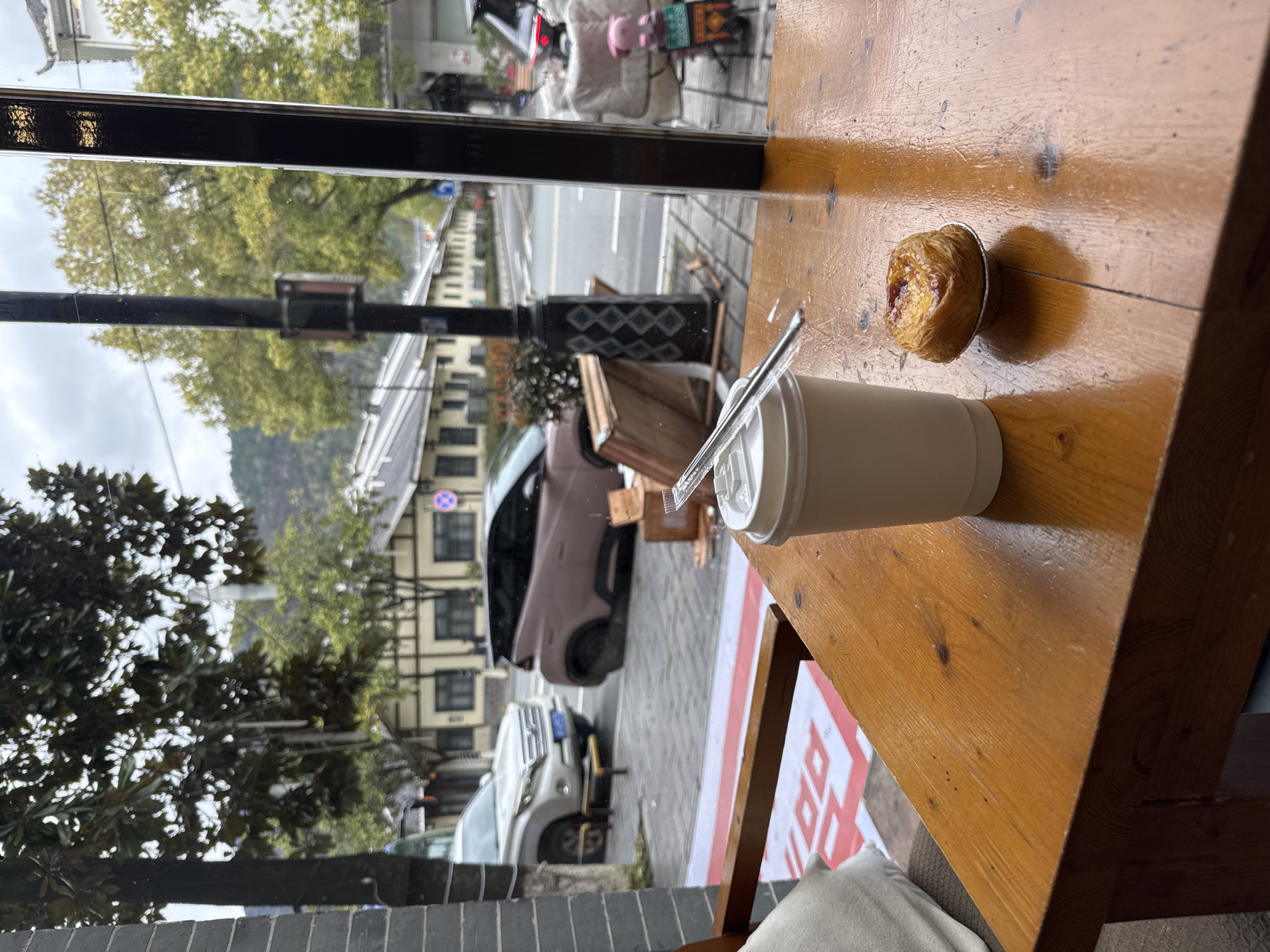 The scene is viewed from inside a establishment, looking out through a large window onto a street in Wulingyuan, China, under an overcast sky. In the immediate foreground, a light brown wooden table surface displays a white disposable coffee cup with a lid and a clear plastic stirrer/straw, positioned to the left. To its right rests a single Portuguese egg tart (pastel de nata) in a small silver foil cup. The wooden table shows visible grain, minor scratches, and natural knots.
Beyond the window, the exterior environment is visible. A grey paved sidewalk and street occupy the midground. A white SUV, resembling a Toyota Land Cruiser Prado, is parked on the left side of the street, facing left, with its front partially obscured by a tree trunk. A yellow metal barrier is positioned directly in front of the SUV. To the right of the SUV, a distinctive two-tone compact electric vehicle, light pinkish-beige with a black roof, is parked facing right. Near this vehicle, several disassembled wooden crates or pallets are stacked haphazardly on the sidewalk. A black utility pole with a decorative diamond pattern is centrally located.
Further down the street on the right, an electric scooter with a pink helmet or seat cover is parked, partially covered by a white sheet or blanket. Buildings line both sides of the street in the background. These are primarily two-story structures with light-colored (cream or beige) facades, dark window frames, and dark, possibly tiled, roofs; some exhibit traditional Chinese architectural elements. A blue circular "no parking" sign with a red diagonal line is visible on the right side of the street. Overhanging green and yellow-leafed trees are abundant, and distant mountains or hills covered in vegetation are visible under the grey sky. No clear human figures are interacting in the immediate or mid-ground, though blurry figures are discernible in the distance. A red and white banner or poster with Chinese characters is partially visible on the ground outside the window, below the parked white SUV.