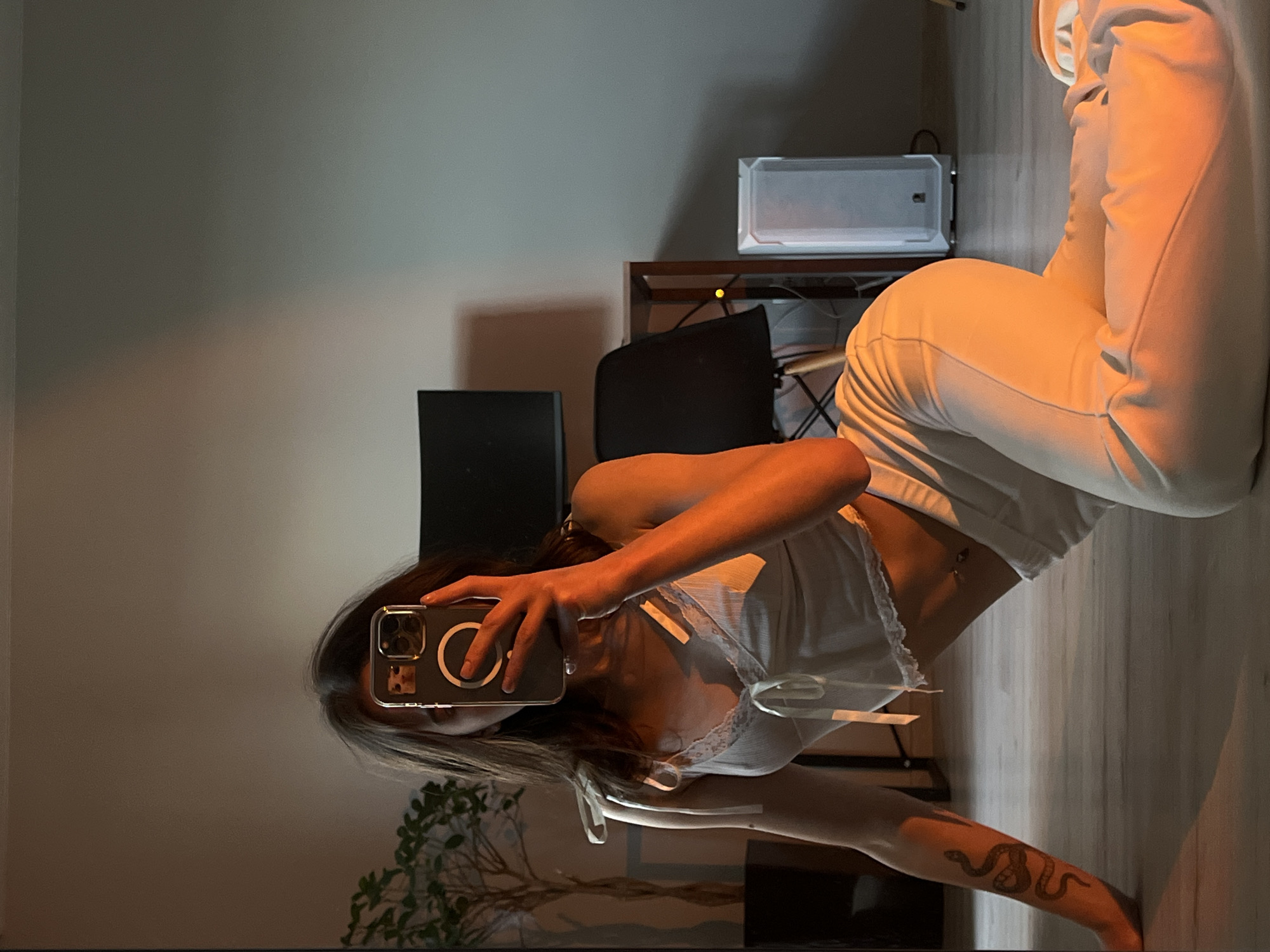 A young woman with dark hair is taking a selfie in a mirror. She is wearing a white crop top and white pants, and has a snake tattoo on her left arm. She is sitting on the floor in a room with a plant, a desk, and a computer tower. The room is lit with warm, orange light. The location is unknown, but it is not Ternopil, Ukraine.