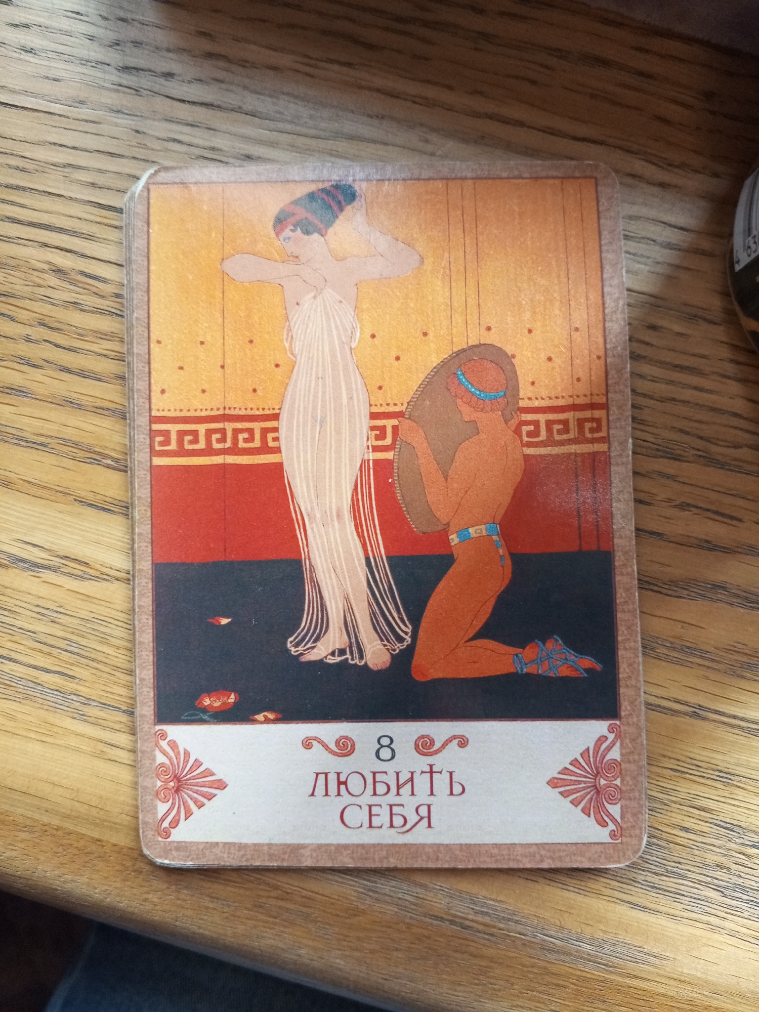 A rectangular oracle or tarot card with rounded corners is centrally positioned on a light brown wooden surface, exhibiting horizontal grain patterns. The card depicts an illustration with a three-tiered background: an upper yellowish-orange section with faint vertical lines and red dots, a middle red band featuring a repeating darker red Greek key (meander) pattern, and a lower dark blue or black section.
On the card, two stylized human figures are presented in an ancient Greek or Roman artistic style. On the left, a slender female figure stands in profile, facing right, with light skin and dark hair styled upwards, secured by a dark band. She wears a long, light-colored, flowing dress with vertical pleats and has her arms raised and bent, appearing to adjust her hair. On the right, a male figure with orange-red skin kneels on his right knee, facing left. He wears a light blue headband and a light blue belt, and blue strappy sandals on his feet. He holds a large, oval, brown shield with both hands, positioned in front of his torso. Several small, red, petal-like objects are scattered on the dark ground between the two figures.
Below the illustration, a light beige band serves as the base for text and decorative elements. The number "8" is visible, followed by the Russian phrase "ЛЮБИТЬ СЕБЯ," which translates to "LOVE YOURSELF" or "TO LOVE ONESELF." Stylized reddish-orange palmette or scroll designs adorn both the left and right corners of this bottom band.
The card is slightly elevated, suggesting a stack of similar cards beneath it. In the upper right corner of the overall image, a portion of a blurred, dark object with a white barcode label is partially visible. At the very bottom edge, a dark blue or black blurred fabric, possibly clothing, is present.
The specific location where this photograph was captured, specifically within the city of Saint Petersburg, Russia, is not discernible from the visual information presented in the image. The setting appears to be an indeterminate indoor environment.