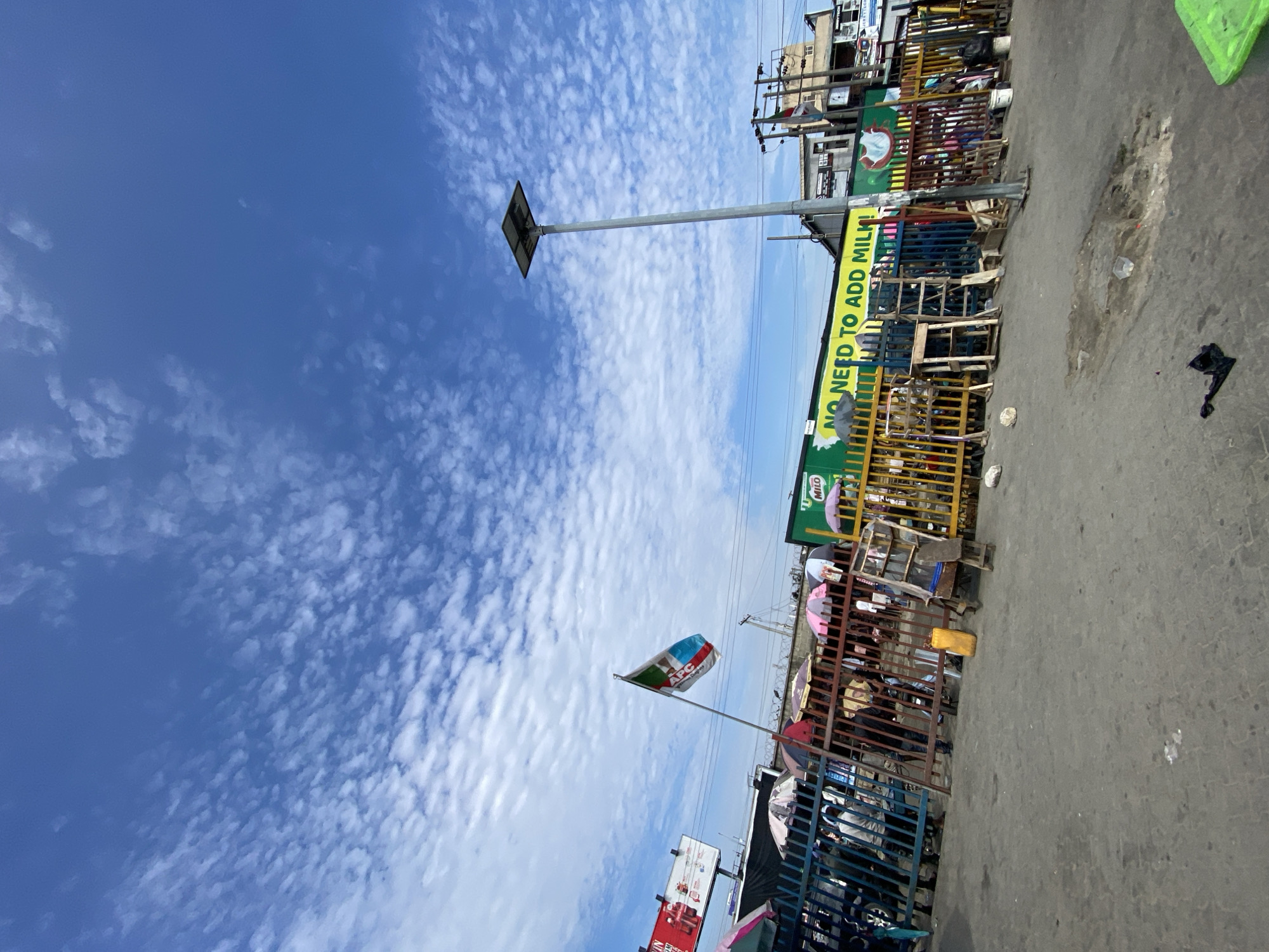 The image shows a street scene in Lekki, Nigeria, under a bright, cloudy sky. The ground is asphalt, showing wear and debris. Along the side of the street are market stalls constructed with metal fencing and supports, housing various goods and vendors. A political flag displaying the "APC" logo is visible. A street light stands tall. Banners advertising "MILO" and "NO NEED TO ADD MILK" can be seen. Overall, the scene depicts a busy marketplace in Lekki.