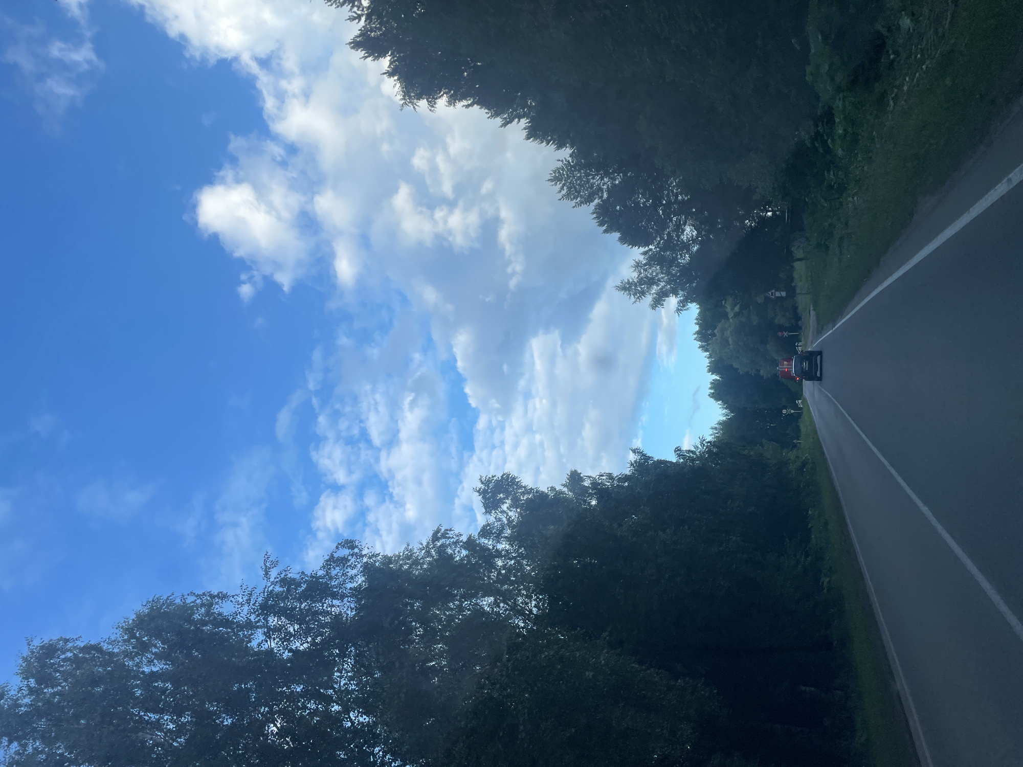 The image captures a view from a vehicle driving on a road surrounded by lush green trees. The sky above is a vibrant blue, dotted with fluffy white clouds. The sun is likely high in the sky, casting a bright light, indicating it's daytime. The road appears to be in good condition, with a white line marking the lane. There is a red vehicle visible ahead on the road, suggesting it is a common occurrence to see other vehicles on this route. The trees lining the road create a tunnel-like effect, framing the sky and the road ahead. The overall impression is one of a peaceful and scenic drive through a natural landscape. There is no visible text in the image. The scene is reminiscent of a summer day in a rural or suburban area.