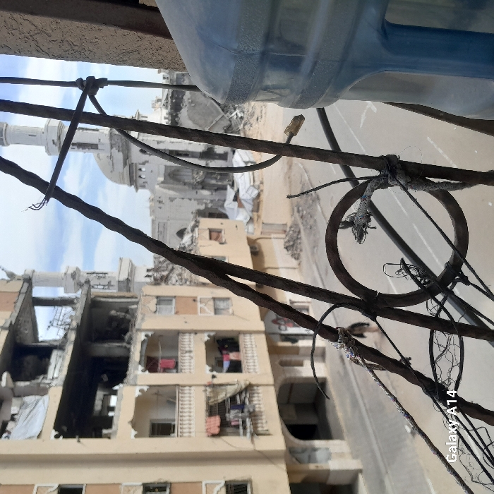 The image presents an urban landscape depicting significant destruction in Khan Yunis, Palestinian Territory, viewed from an elevated perspective. In the immediate foreground, rusty metal bars, possibly part of a railing, are prominently featured, entwined with various wires and ropes. A large, translucent blue plastic water dispenser bottle is partially visible on the far right.
Across a paved street, a multi-story building on the left shows extensive structural damage on its upper floors, with collapsed sections exposing interiors and rebar. Lower floors of this building feature balconies with clothes hanging to dry. A solitary, dark silhouette of a person is visible walking on the street in the midground. Piles of rubble and debris are scattered along the street and surrounding areas, with several white tarpaulins or makeshift shelters visible amidst the wreckage further down the street. Dominating the background is a severely damaged mosque, characterized by a large dome and at least one partially intact minaret, with large sections of its facade and structure destroyed. The sky is partly cloudy.