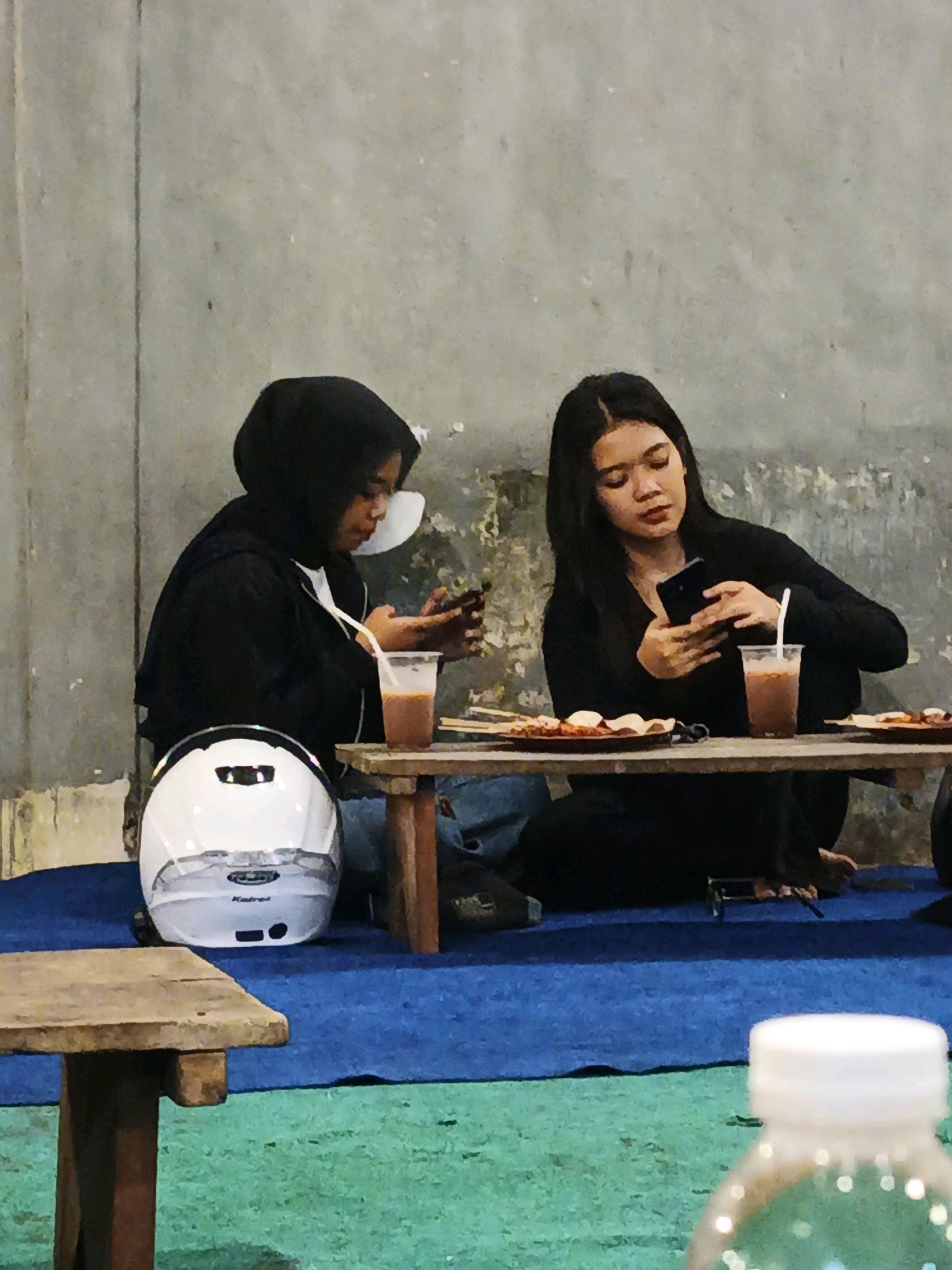 Two young women are seated on the floor at a low table, which is set with food and drinks. They are both looking at their mobile phones. The woman on the left is wearing a black hijab and a black jacket over a white shirt, and she has a plastic cup with a straw in front of her. A white motorcycle helmet is to her left. The woman on the right is wearing a black top and has long dark hair. She has a plastic cup with a straw and a plate with food in front of her. The background is a plain concrete wall. The scene appears to be an outdoor setting, possibly a food stall or informal dining area, in Tulangan, Indonesia. The ground is covered with a blue mat, and a wooden table is in the foreground.