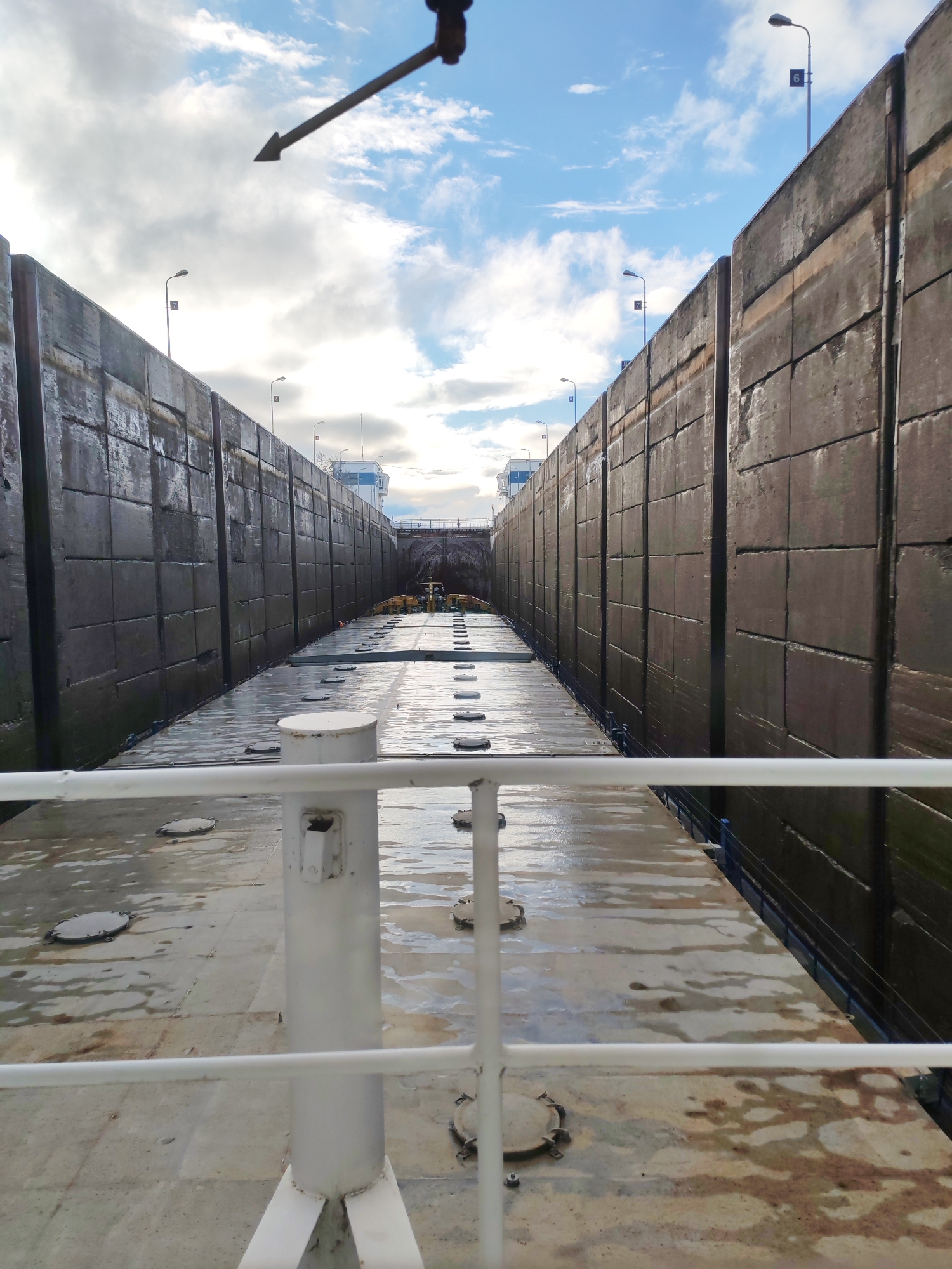 The image depicts the view from a vessel inside a ship lock, likely on a canal or river. Two massive concrete walls, segmented into large blocks, rise on either side of the vessel. Light poles with number plates are visible atop the walls, indicating numbered sections of the lock.
In the foreground, a white metal railing partially obscures the view. The deck of the vessel is wet and shows circular access hatches. Further ahead, additional equipment is visible. A control tower structure is present between the walls at the far end of the lock.
The sky above is partly cloudy, with patches of blue visible. A dark, arrow-shaped object intrudes from the top center of the frame, its origin unclear. The location is unknown, and no people are clearly visible.