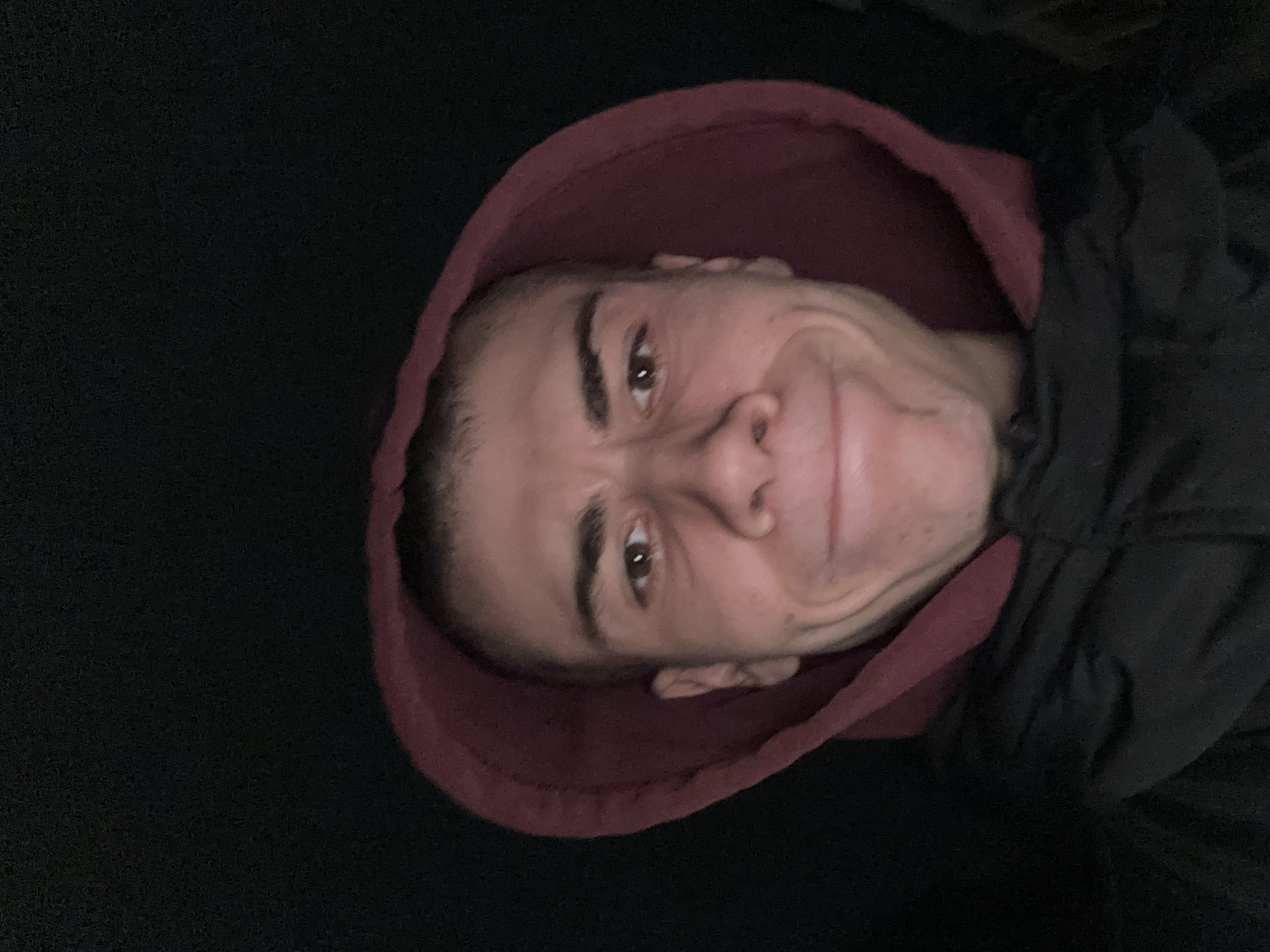 A rotated close-up photograph features a person's face centered in the frame. The person has dark hair, a fair complexion, and is wearing a red hooded sweatshirt and a black jacket. The background is a dark solid color. The location is not identifiable from the image.