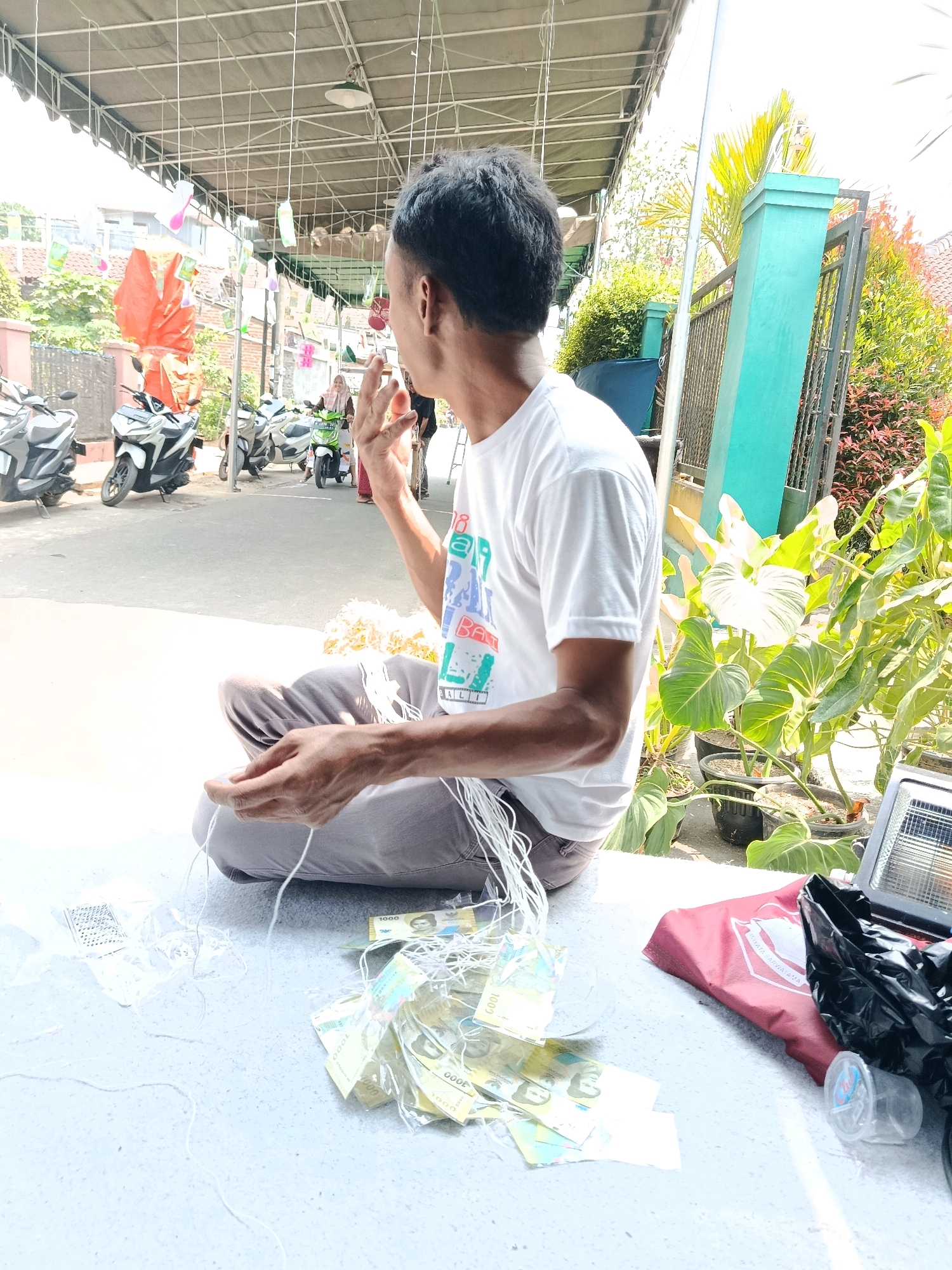 A man sits cross-legged on a white surface, facing to the right, in what appears to be an outdoor marketplace in Bululawang, Indonesia. He wears a white t-shirt and gray pants. His right hand is raised to his face, and he appears to be smoking a cigarette. In front of him, on the white surface, are several strands of white string, some of which are attached to small, transparent plastic bags containing Indonesian Rupiah banknotes. A pile of similar banknotes is also visible on the surface. To the right of the man, there are potted plants, a red bag, a black plastic bag, and a small light fixture with a metal grate. In the background, several motorcycles are parked, and a covered walkway extends into the distance. The overall scene suggests a commercial or craft-making environment.