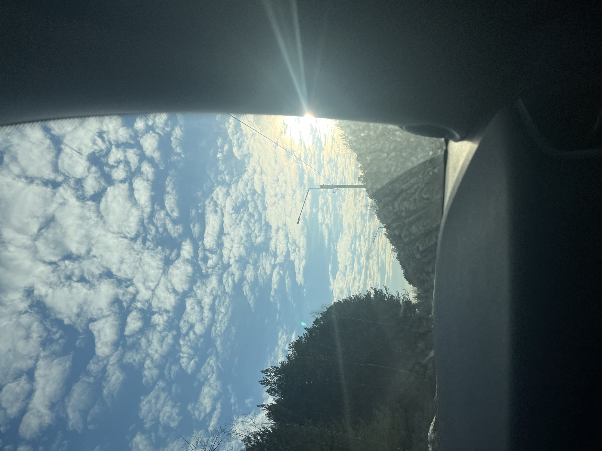 The image shows a view from inside a vehicle, looking outwards through the windshield. The sky is visible, featuring a combination of blue sky and white, puffy clouds. The sun is shining brightly, causing a glare near the top of the windshield. In the distance, there are trees visible, along with a utility pole. The interior of the vehicle is visible, with parts of the dashboard and ceiling. The exact location in Chaikovsky, Russia is not determinable from the image without further location details.