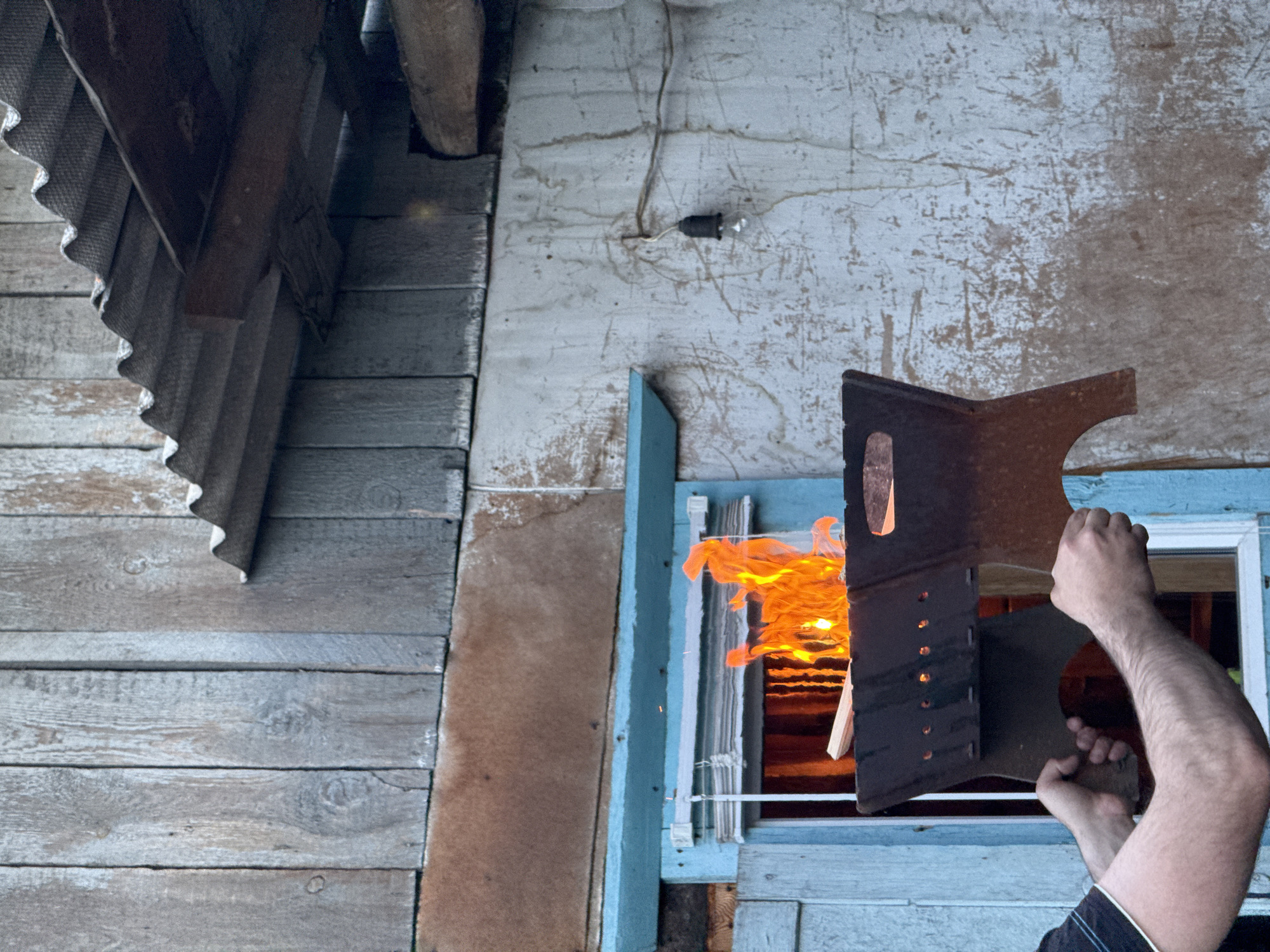 The image shows a person holding a metal object in front of a window with flames visible inside. The exterior of the building is wooden, with a weathered or peeling paint finish. The location is potentially in Зыково, Russia, based on provided information. The metal object has a geometric design with holes and cutouts. The person's arms are visible, but other details are obscured. The environment appears to be an older, possibly rural or suburban, setting.