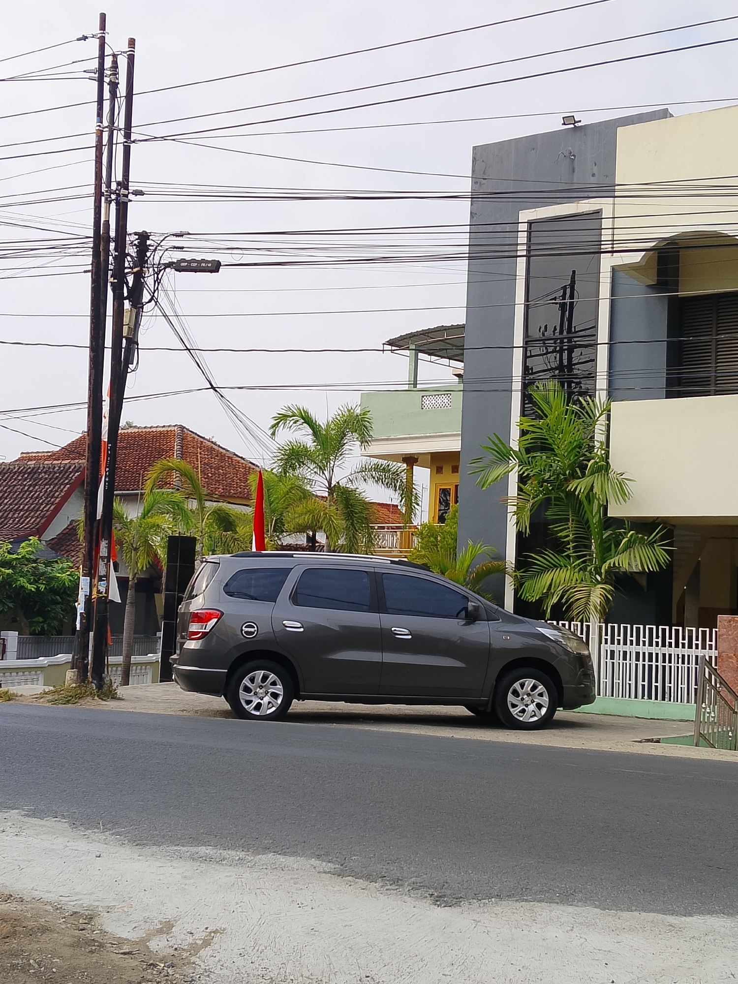 A grey minivan is parked on the side of a paved road in the city of Ngelo, Indonesia. The minivan is facing right, with its rear end closer to the viewer. A modern building with a grey and cream facade is situated to the right of the minivan, with a white fence in front of it. To the left of the minivan, there are palm trees and a residential building with a tiled roof. Utility poles with numerous wires are visible in the background. The sky is overcast. There are no people visible in the image.