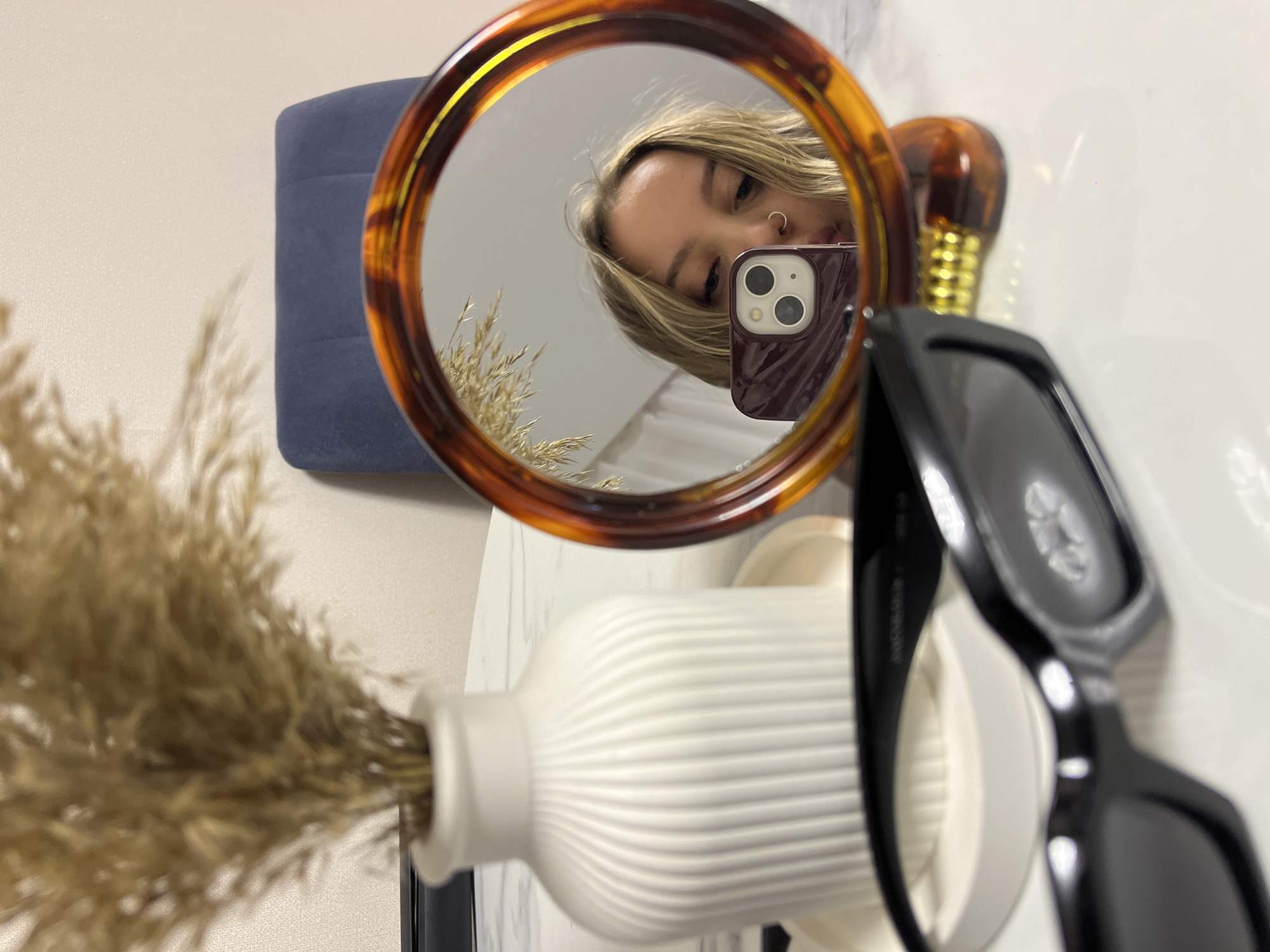 This media file depicts an indoor scene in Krasnodar, Russia, featuring a mirror selfie. The central subject is a young woman, partially visible in the reflection of a prominent circular mirror with a tortoiseshell-patterned frame. She has blonde hair and a nose piercing, and her eyes are directed towards the smartphone she holds, which has a dark red case and a triple-camera setup. Her face shows a neutral expression.
Surrounding the mirror are various objects. To the left, a substantial bundle of dried ornamental grass, likely pampas grass, is arranged in a white ceramic vase with a ribbed texture. A reflection of this grass is also visible within the mirror. To the right of the mirror, a decorative light bulb with a clear glass globe and a golden-colored base is partially seen, blurred. A pair of black sunglasses rests on a surface in the foreground on the right, also somewhat out of focus. In the background, a dark blue or navy upholstered item, possibly a cushion or part of a furniture piece, provides a soft backdrop. The setting suggests a styled interior space, potentially a vanity or living area. The time of day is not discernible, and no text is visible in the image.