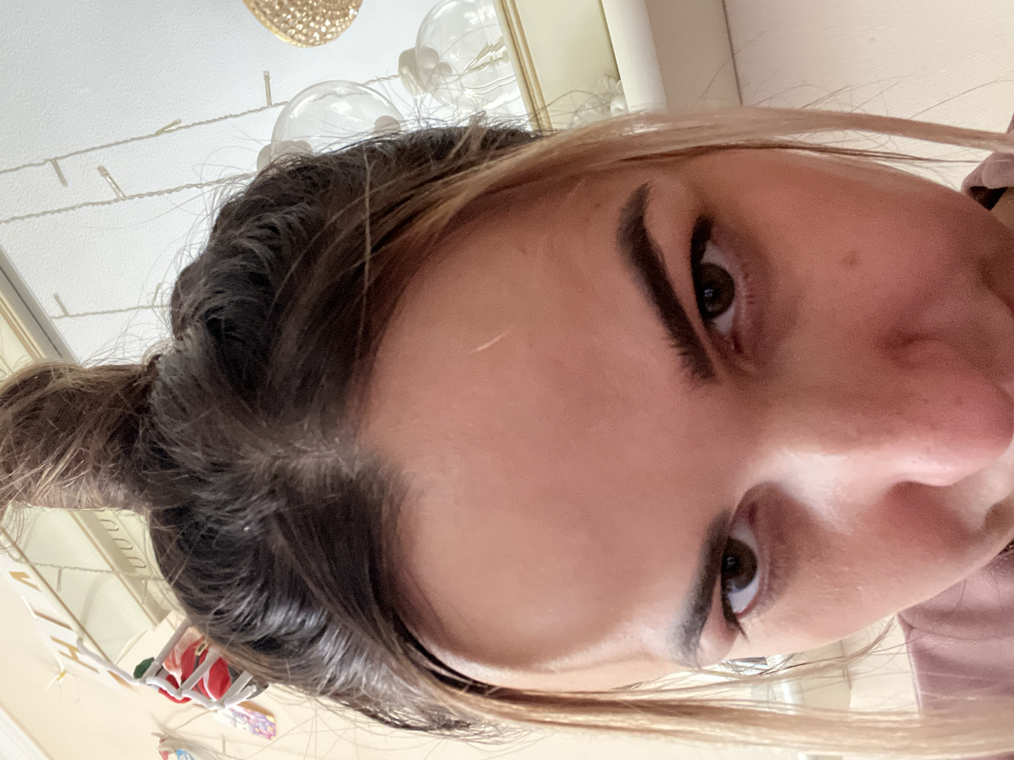 The image is a close-up, overhead, selfie-style shot of a person's face, with their head tilted down. The person has dark brown hair pulled up into a bun, with lighter brown strands framing their forehead. Their eyes are dark brown and appear to be looking directly at the camera. The lighting is bright, casting shadows on the face. In the background, there are reflections in a mirror and some decorative lights, possibly holiday-themed. The setting appears to be indoors.