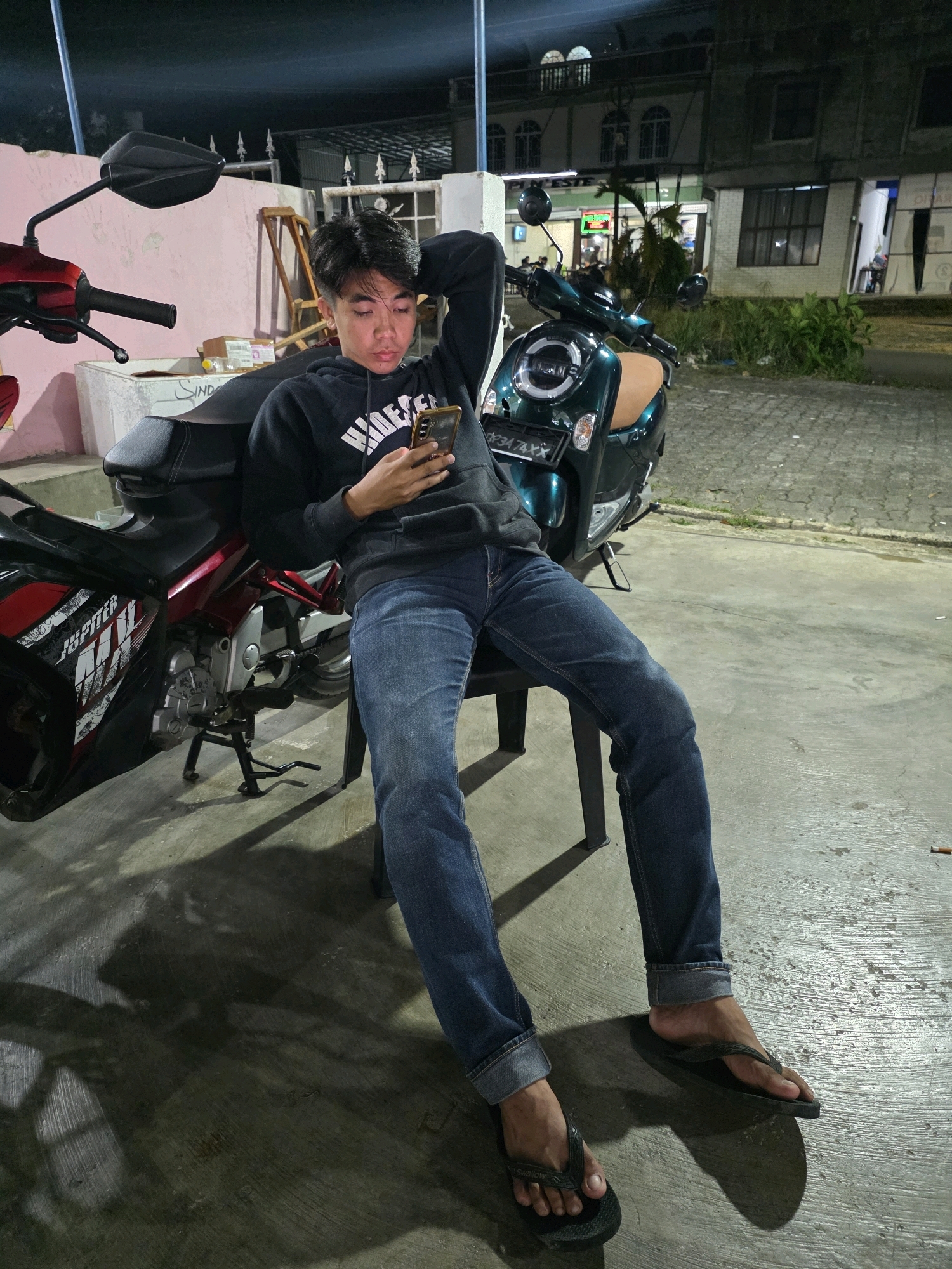 In Tanjung Pinang, Indonesia, a young man in jeans and a black hoodie sits on a chair next to a red and black motorcycle. He is looking at his smartphone, with one hand on the phone and the other resting on his head. Behind him, a dark green scooter is parked. The scene appears to be outdoors at night, with artificial lighting illuminating the area. Buildings and vegetation are visible in the background. The ground is a concrete surface with shadows cast by the motorcycle and the man.
